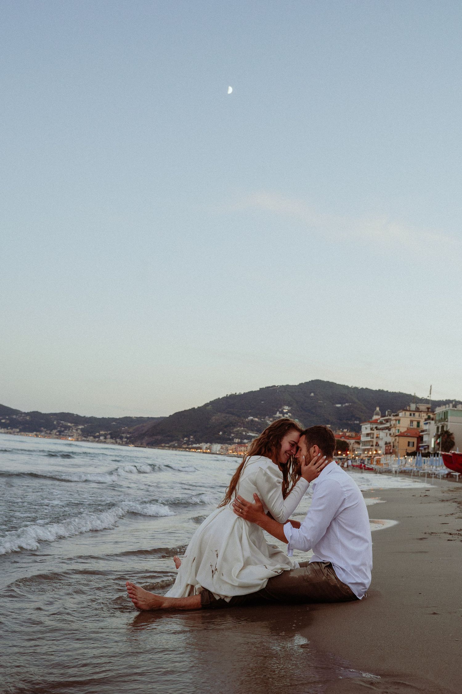 Couple sitting close together on wet sand sharing a kiss with crescent moon over coastal town at dusk.