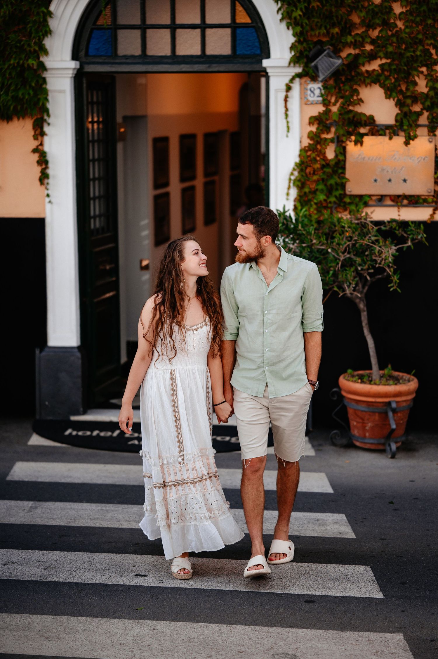 Happy couple holding hands and smiling while crossing a zebra crossing outside a charming hotel entrance.