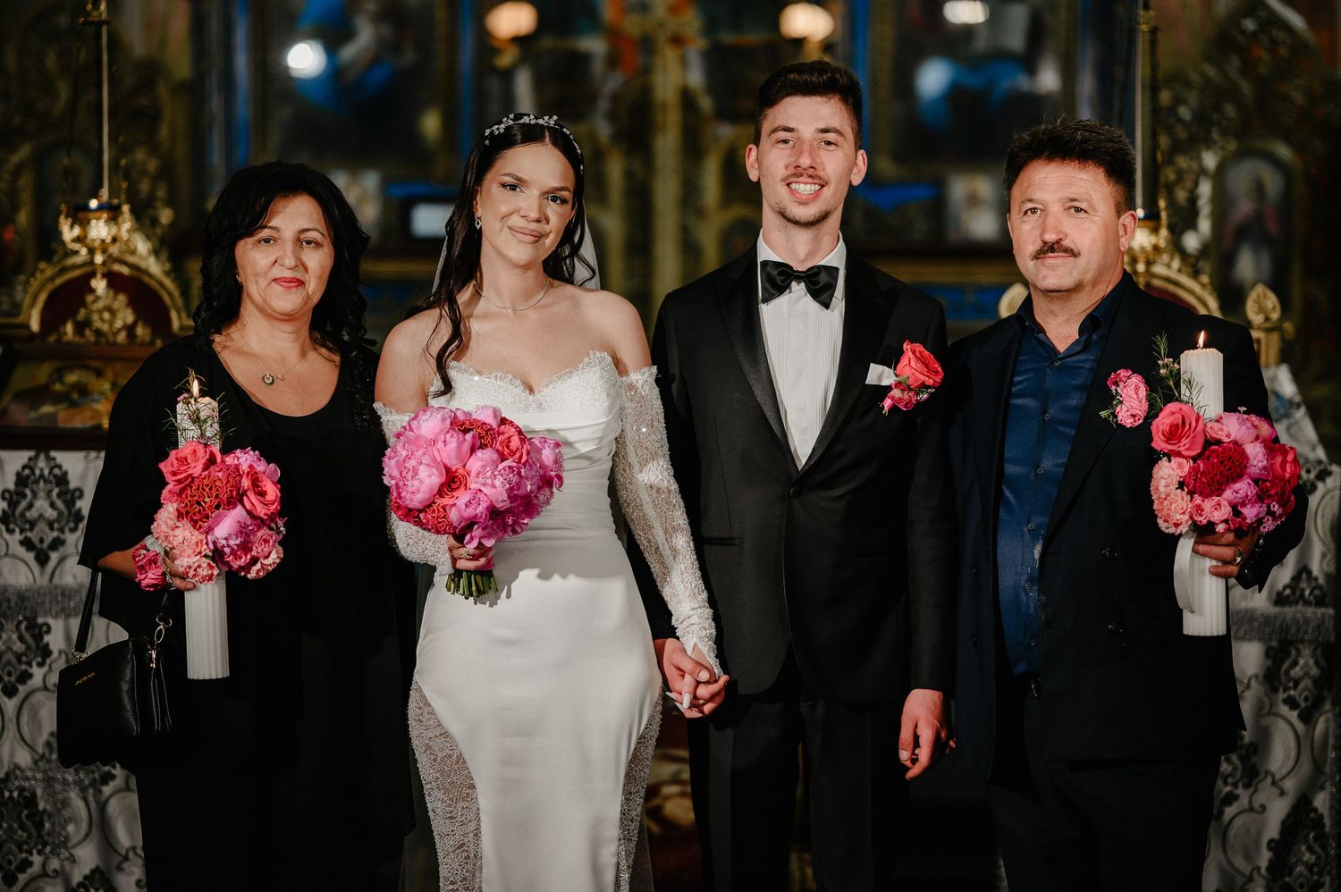 Bride and groom pose with parents holding pink peony bouquets inside a decorated Orthodox church.