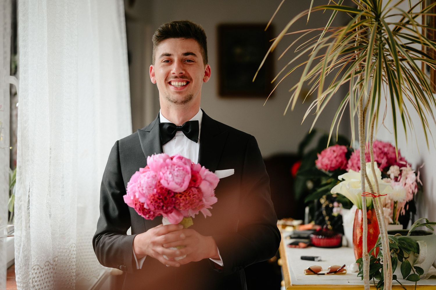 Smiling groom in black tuxedo holds pink peony bouquet near window on wedding day.