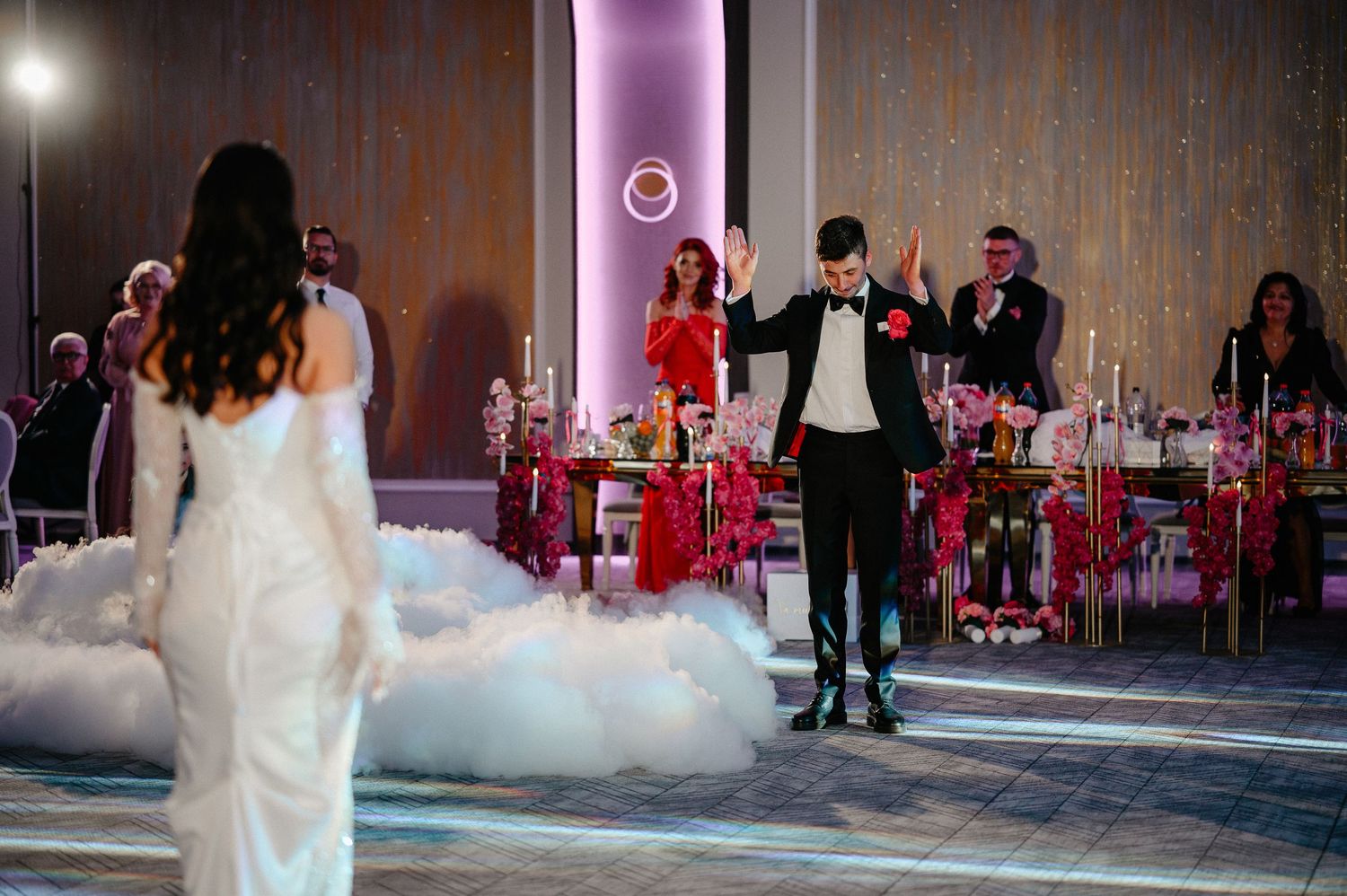 Groom in tuxedo gestures dramatically to bride on fog-covered dance floor at glamorous wedding reception.
