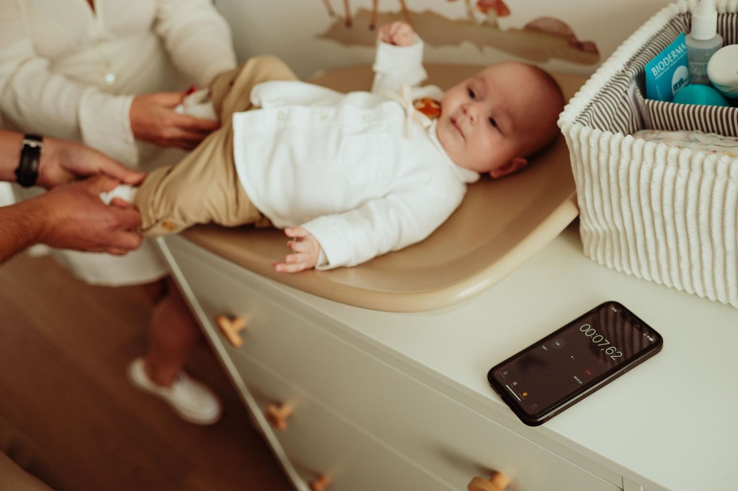 Newborn baby in white outfit lying on changing table with smartphone nearby.