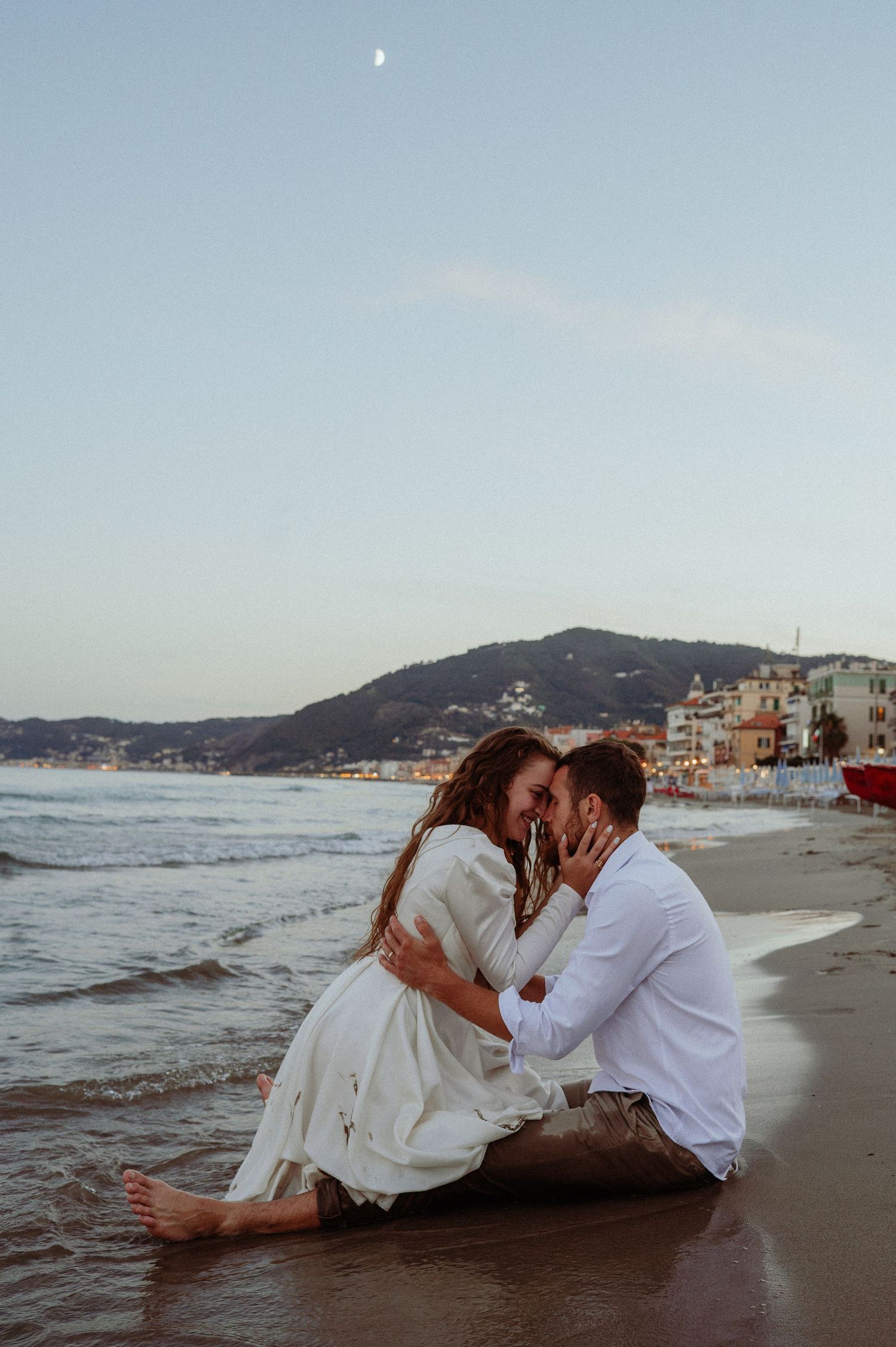 Romantic couple sitting on sandy beach at sunset, embracing and smiling near ocean waves.