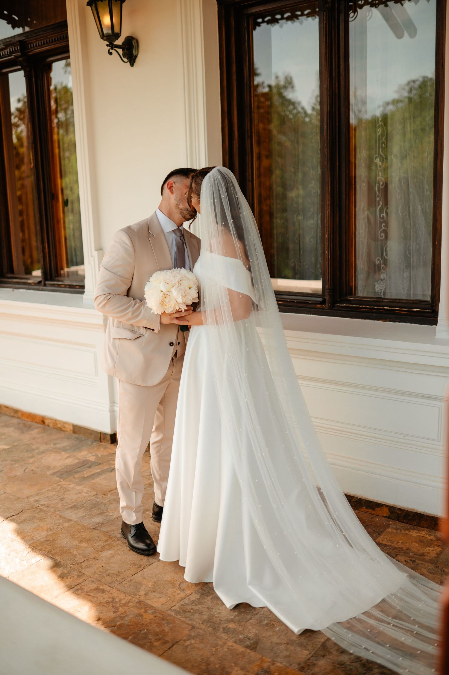 Bride and groom share a kiss on sunlit porch, bride in white flowing gown with veil holding white floral bouquet.