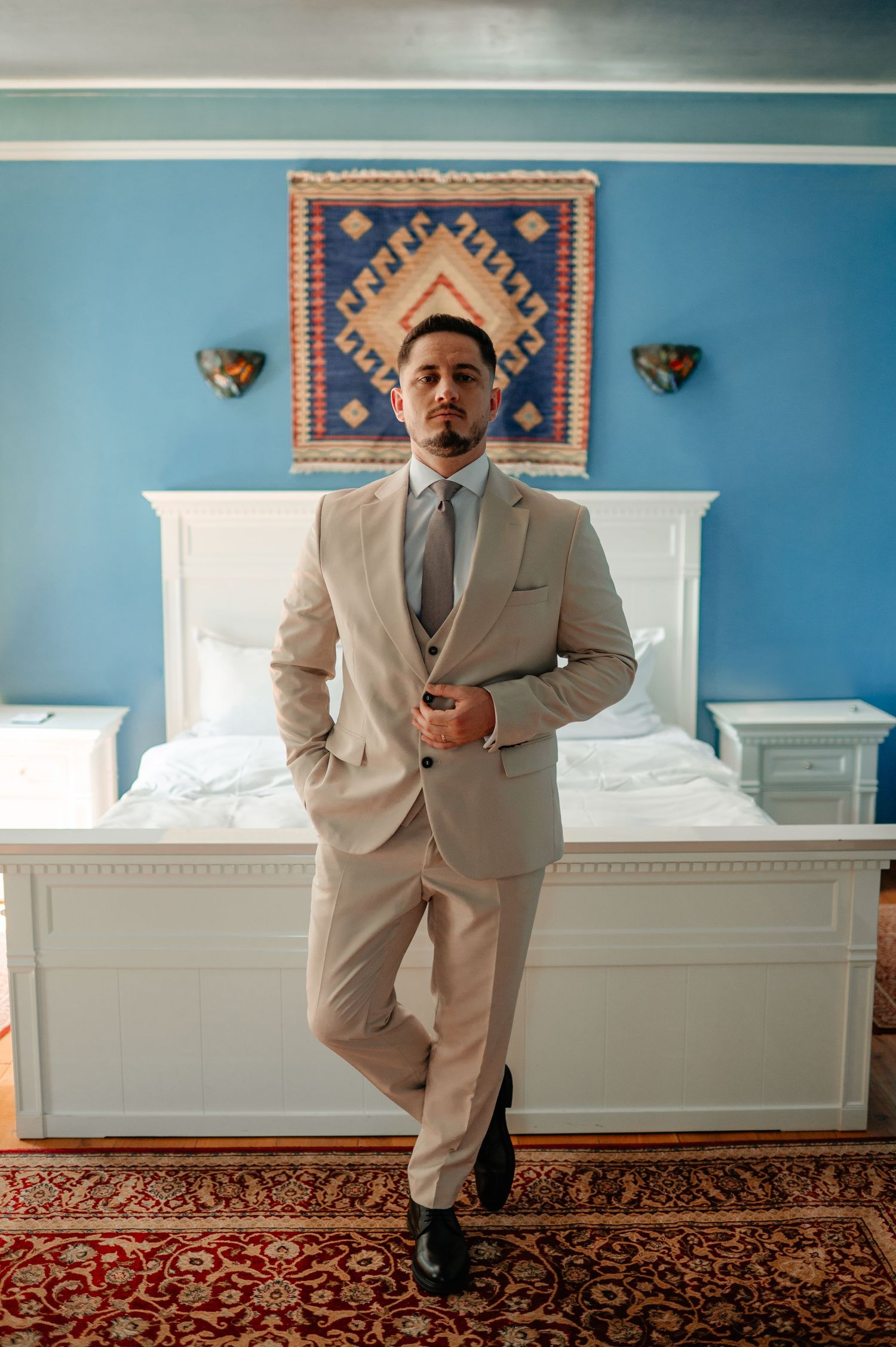 Dapper groom in a beige three-piece suit posing confidently in a blue bedroom before his wedding.