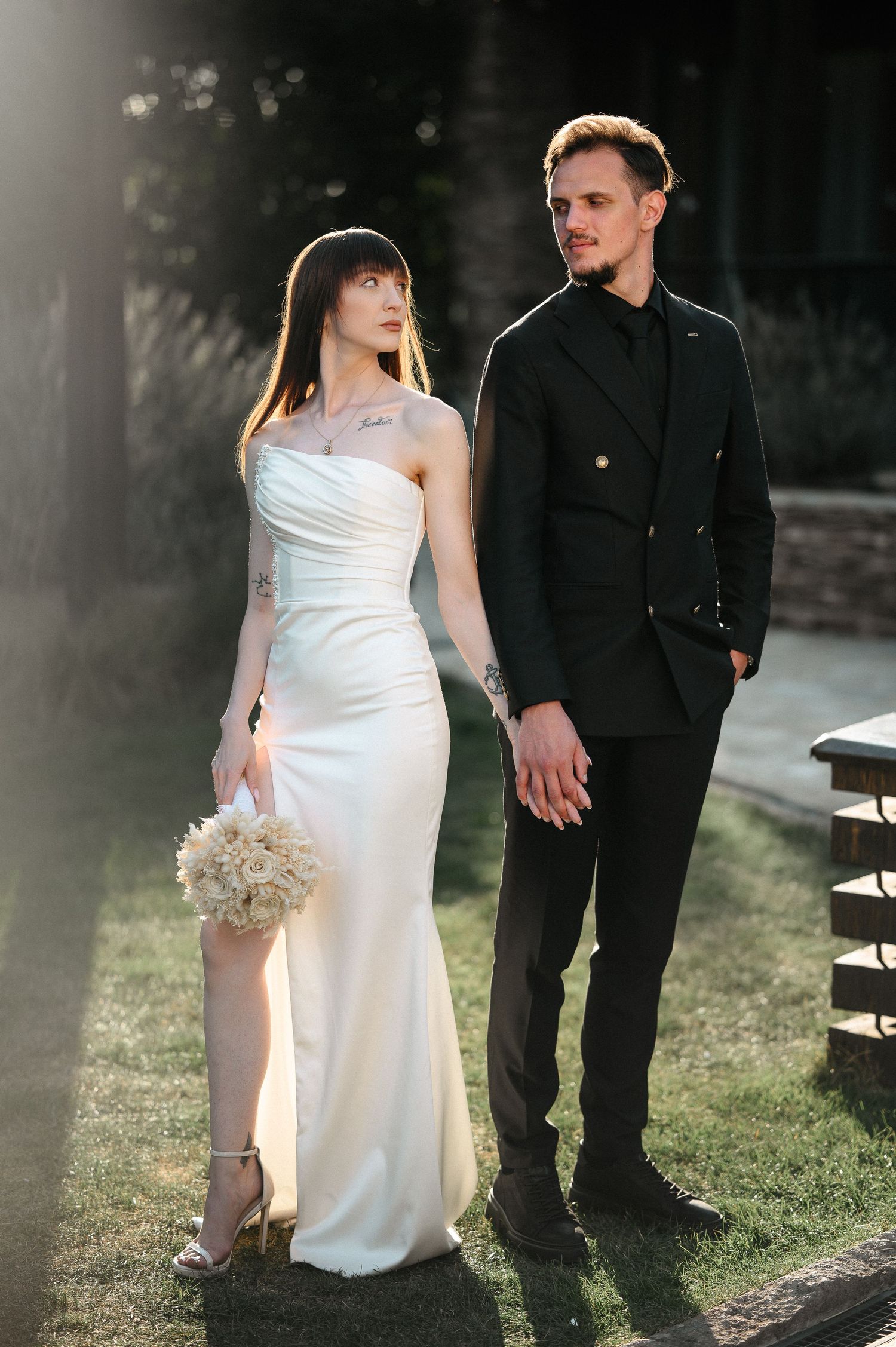 Bride and groom holding hands in sunlit garden, she in white slit gown, he in black double-breasted suit.