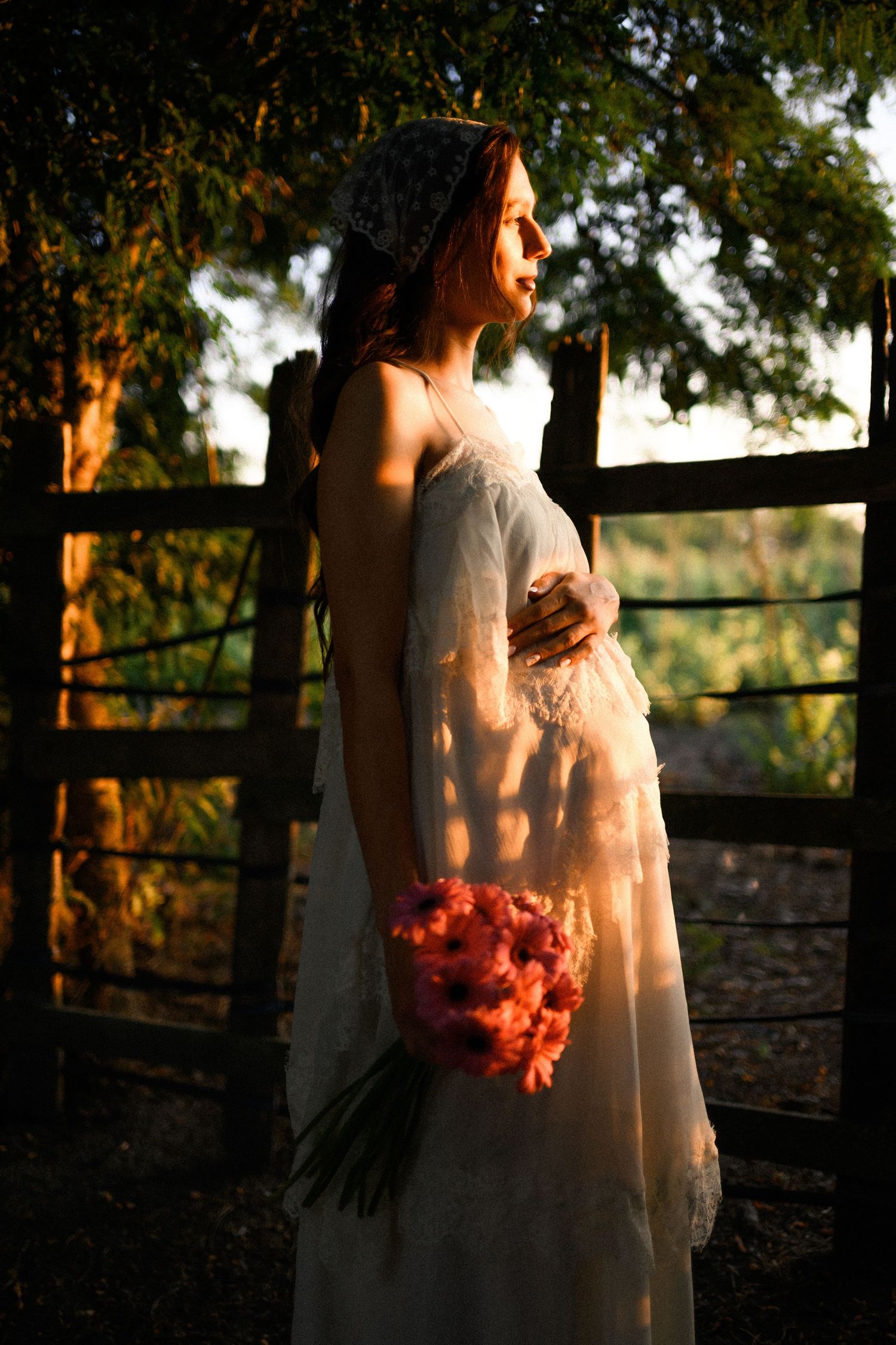 Pregnant woman in white lace dress holding pink flowers at golden hour by a rustic wooden fence.