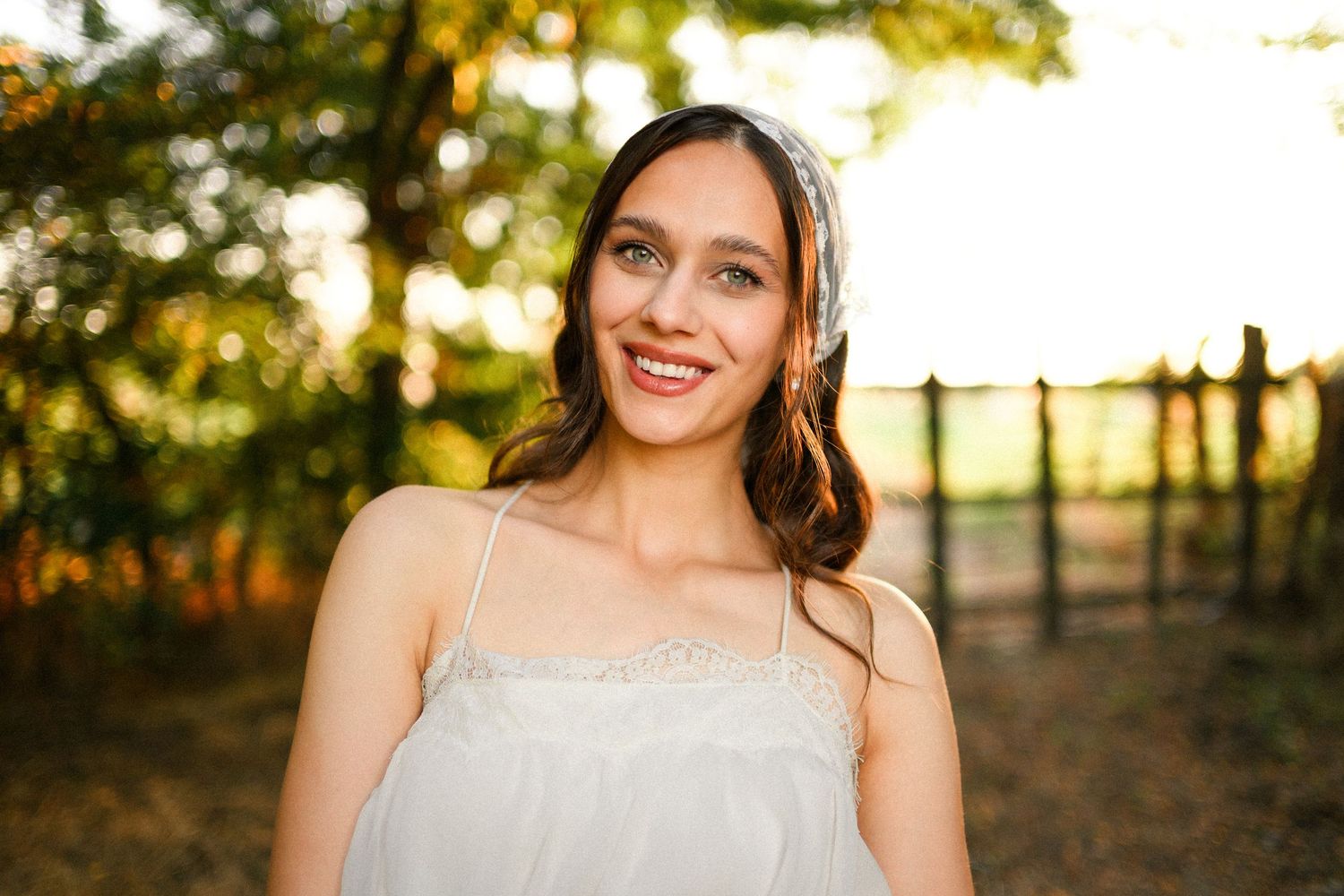 Smiling young woman in white sundress with headband poses outdoors in golden hour sunlight near wooden fence.