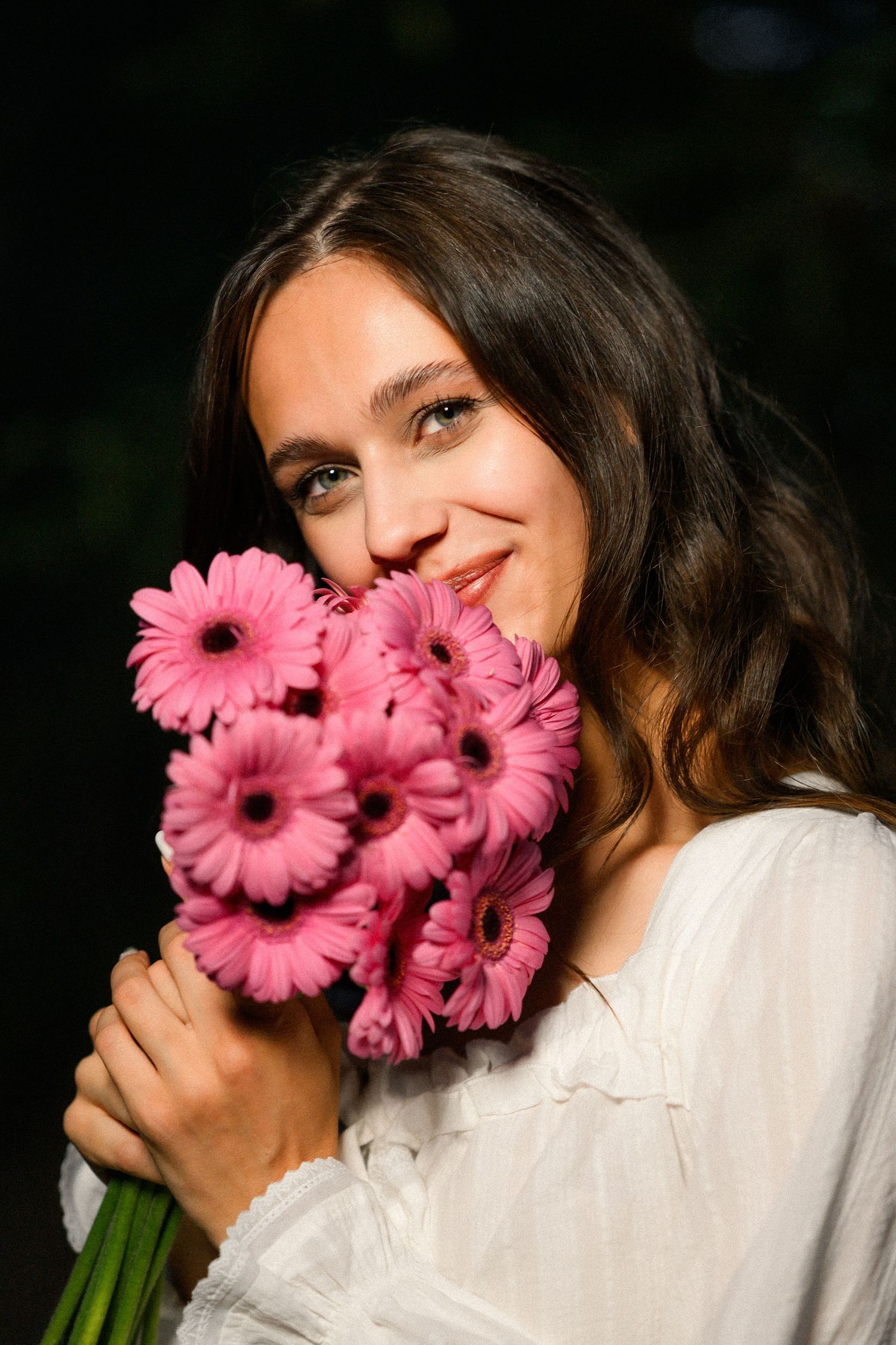 Young woman with dark hair holding a bouquet of pink gerbera daisies, smiling softly in natural light.