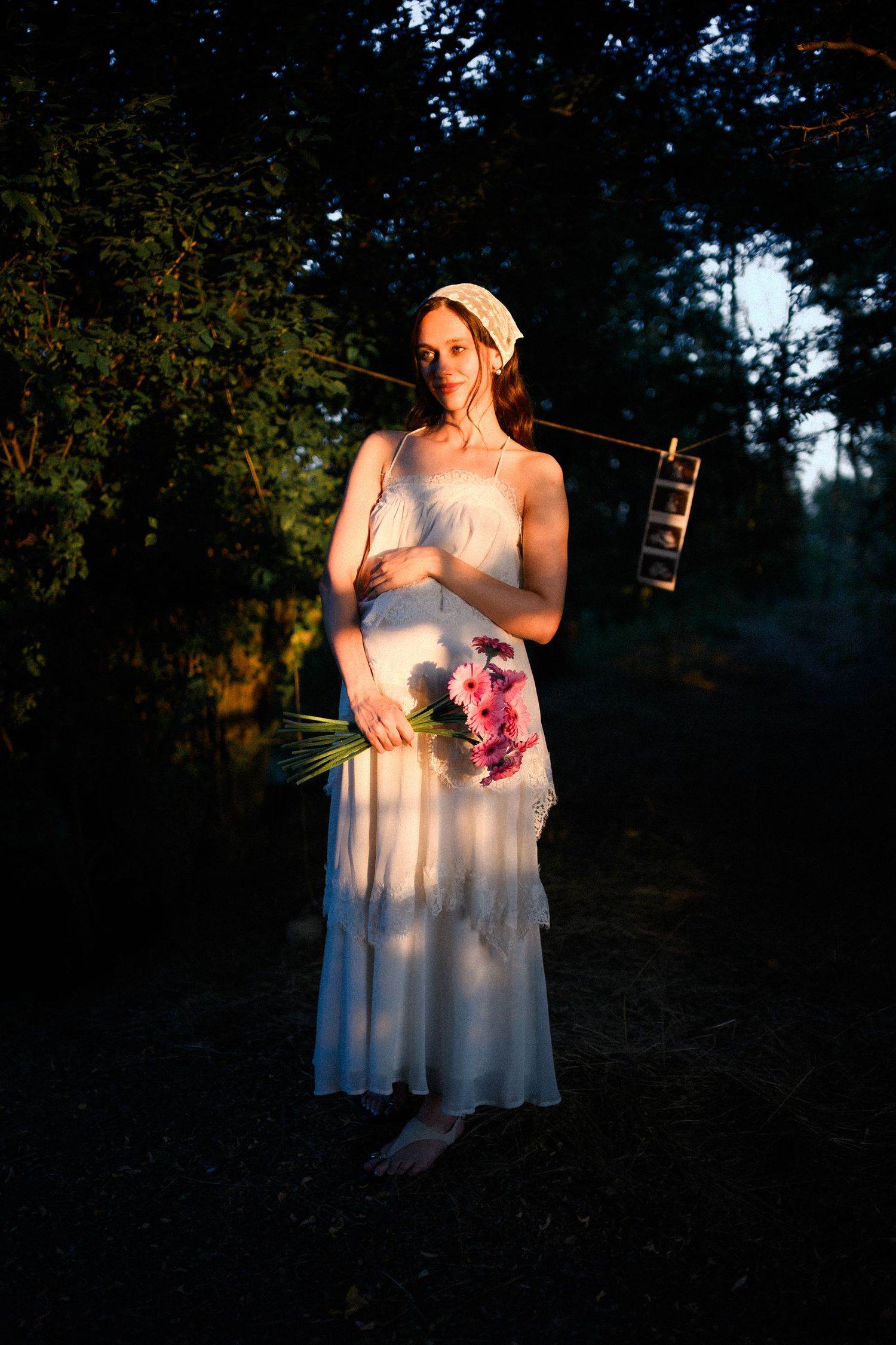 Woman in white dress holding pink flowers, bathed in golden sunset light in a dark forest setting.