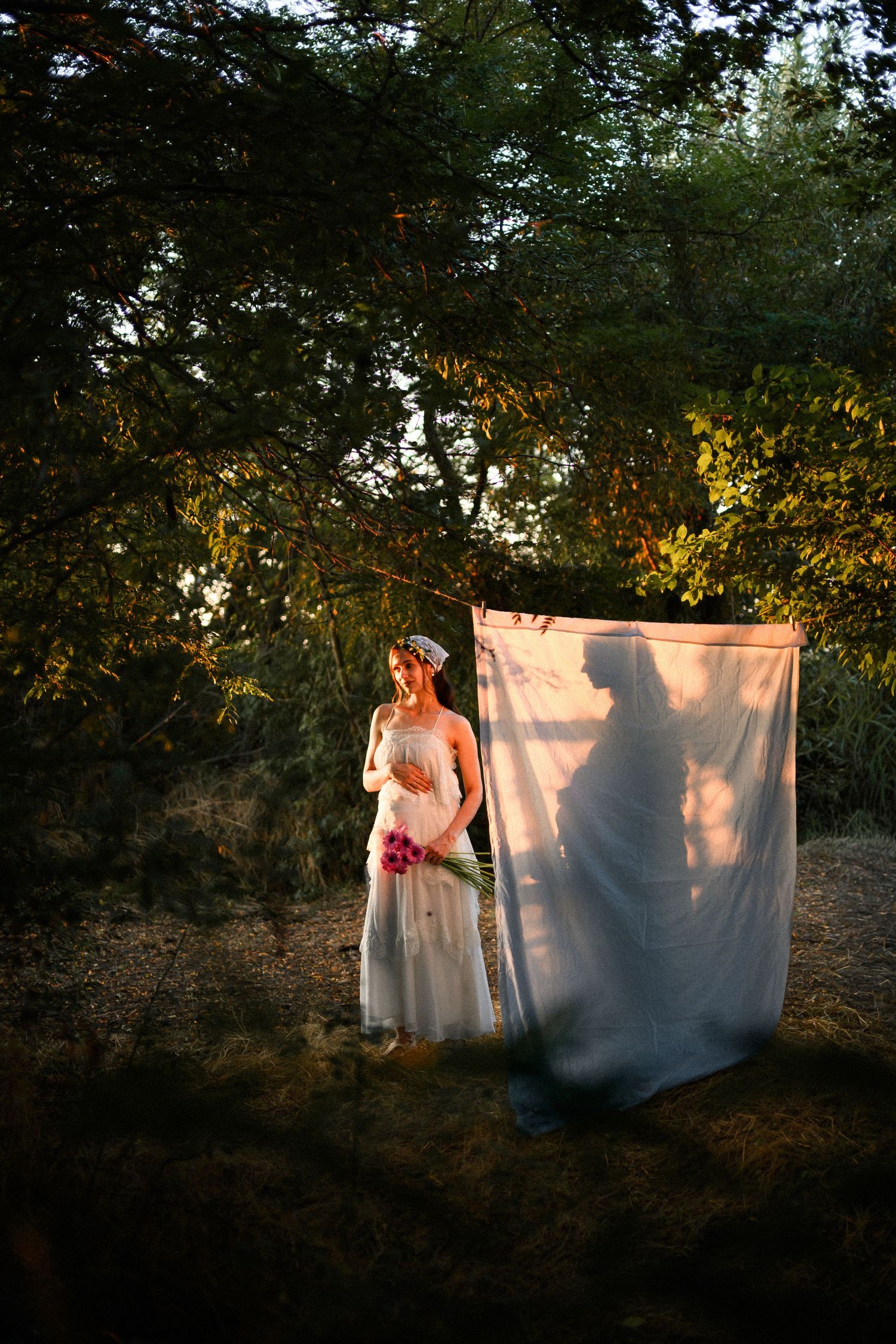 Bride in white gown holding pink bouquet stands beside draped fabric with silhouette shadow in forest setting.