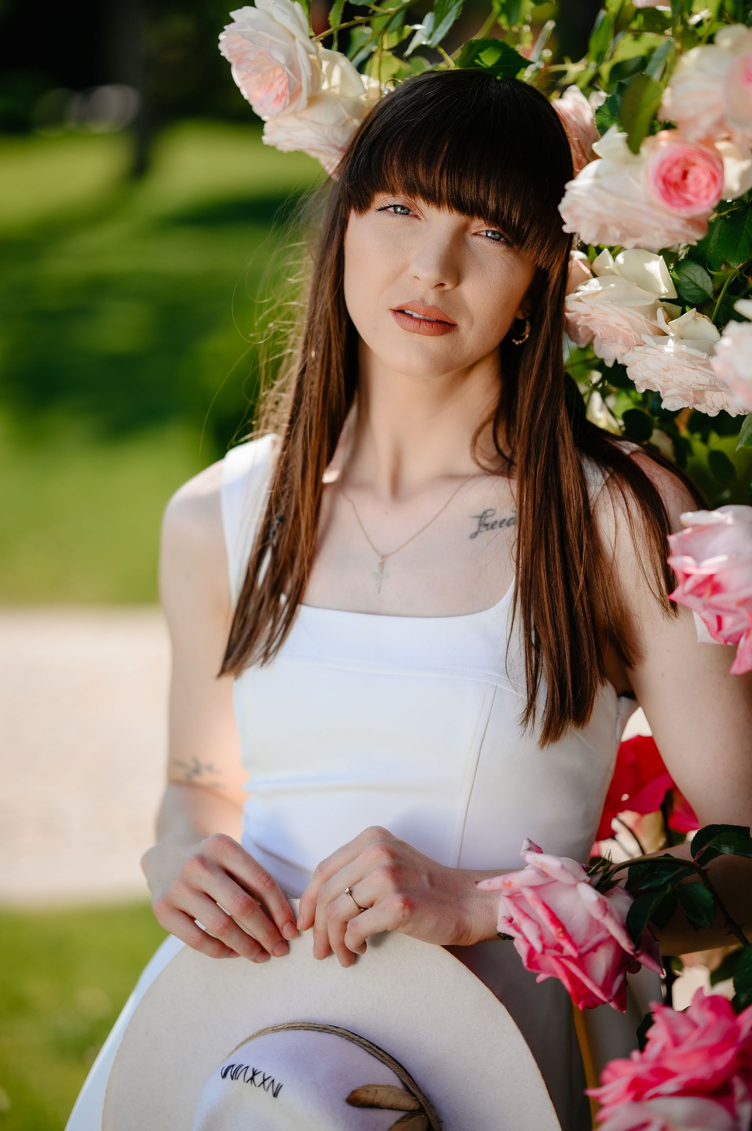 Young brunette woman in white dress holding a hat poses beside blooming pink roses in a sunny garden.