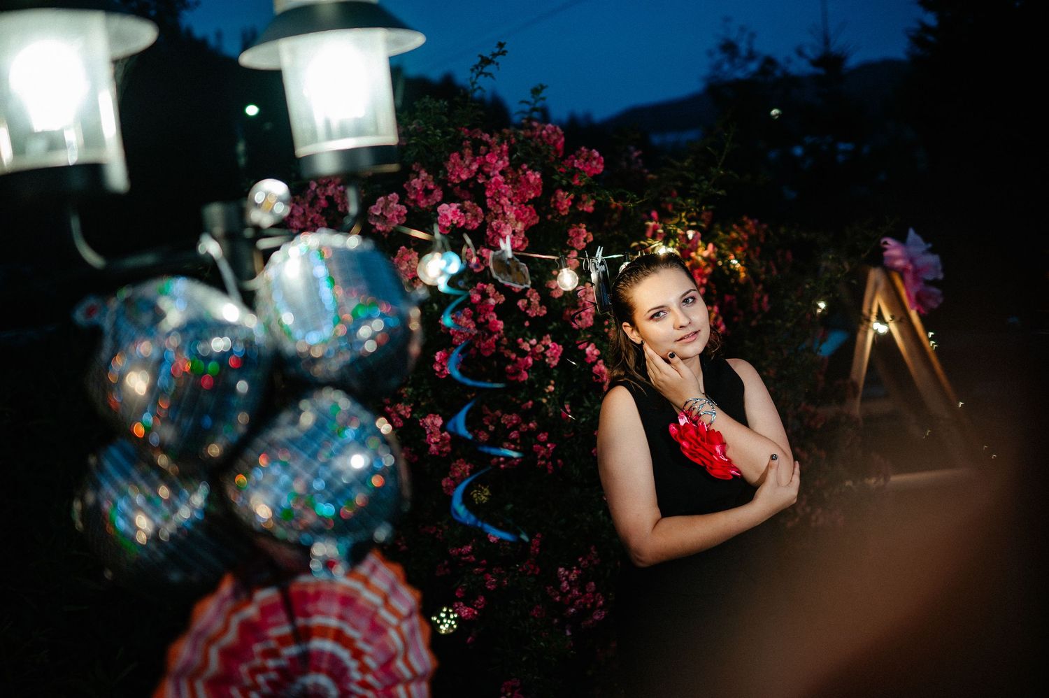 Young woman in black dress smiles among pink flowers and disco ball decorations at night party.