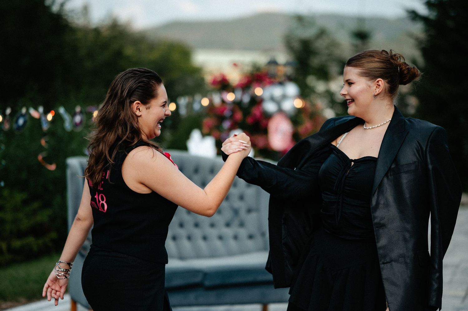 Two women in black dresses laughing and holding hands at an outdoor evening party.