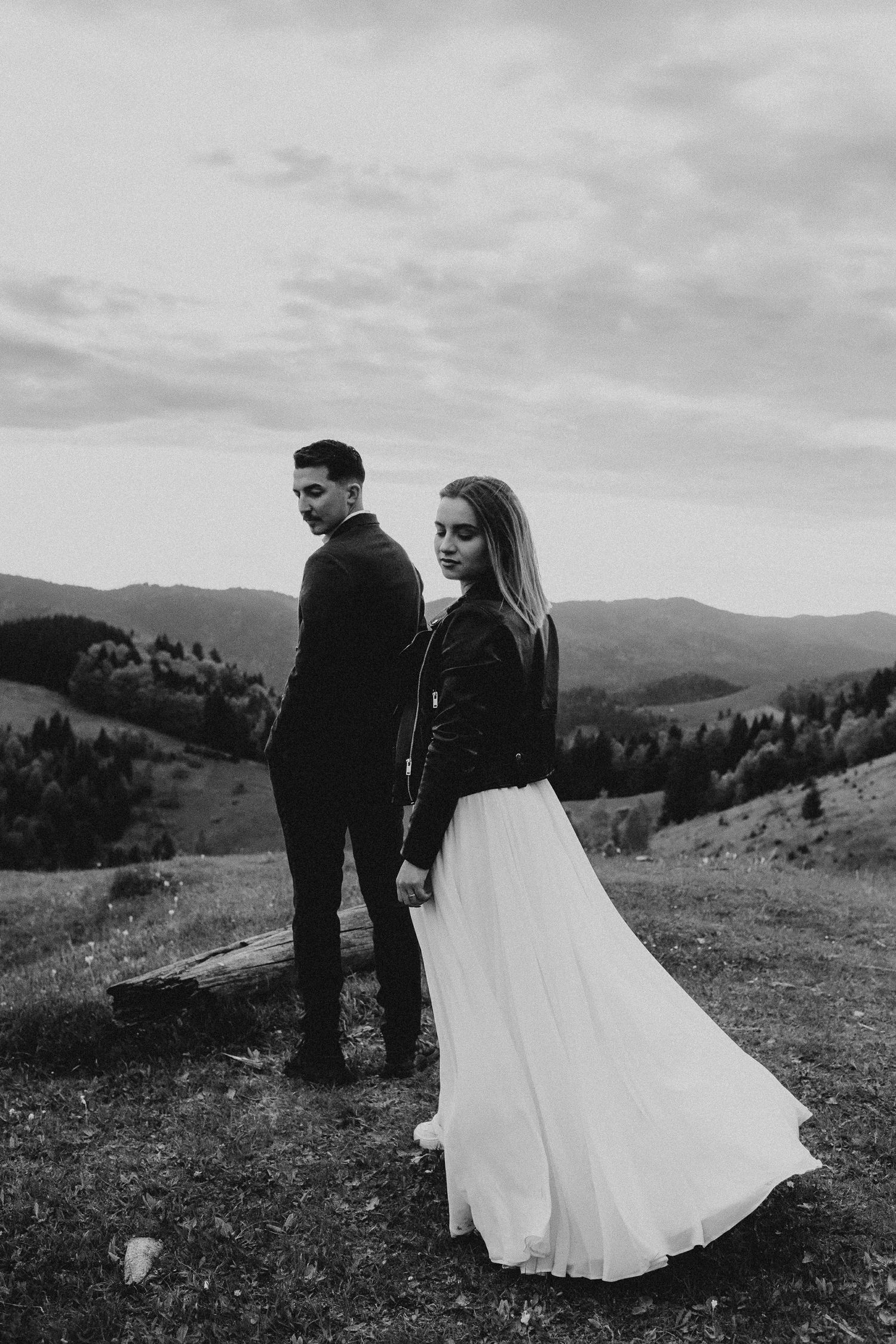Bride in white skirt and leather jacket stands with groom on scenic mountain hillside in dramatic black and white.