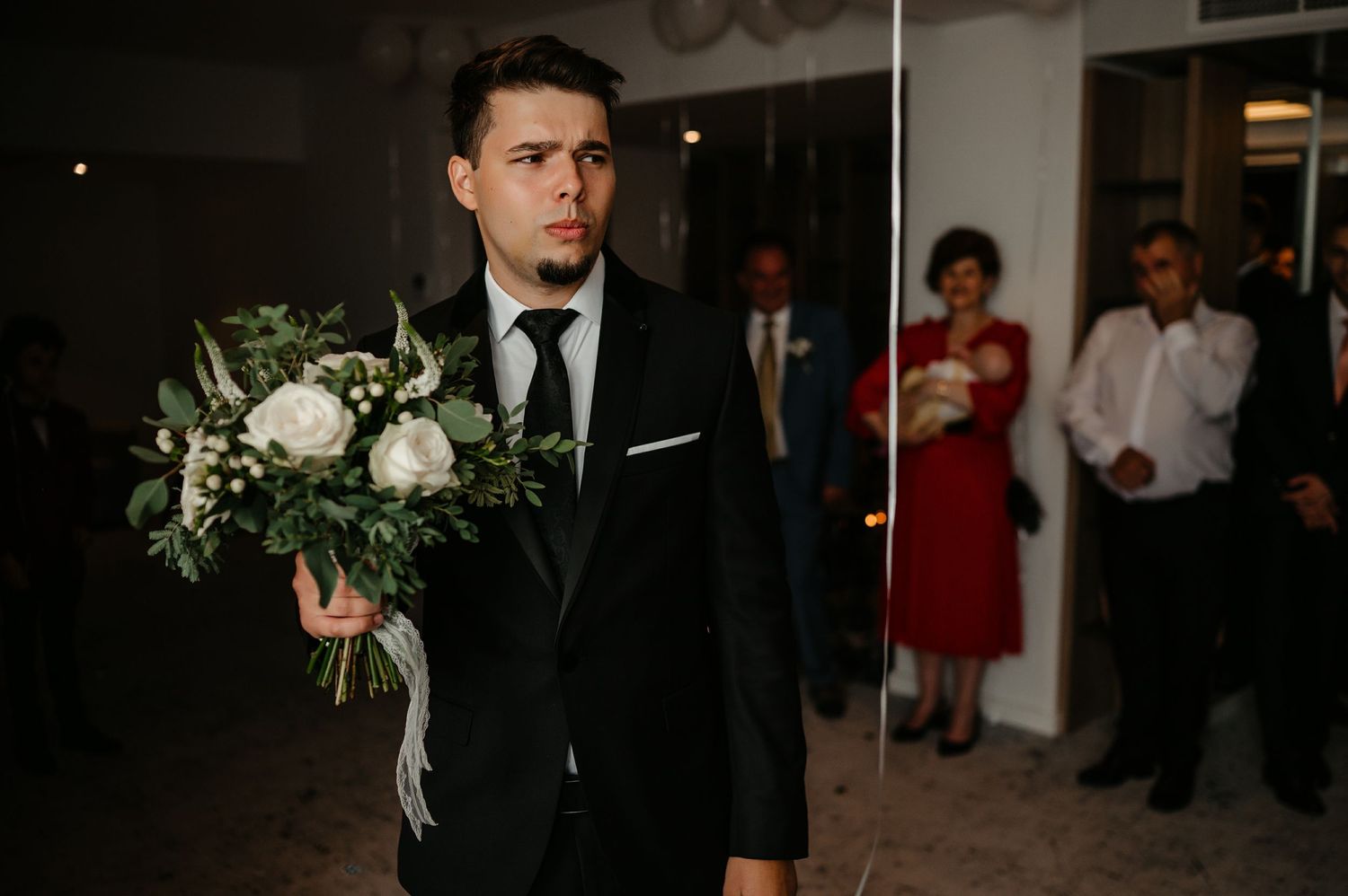 Serious groom in black suit holding white floral bouquet with guests watching in background.