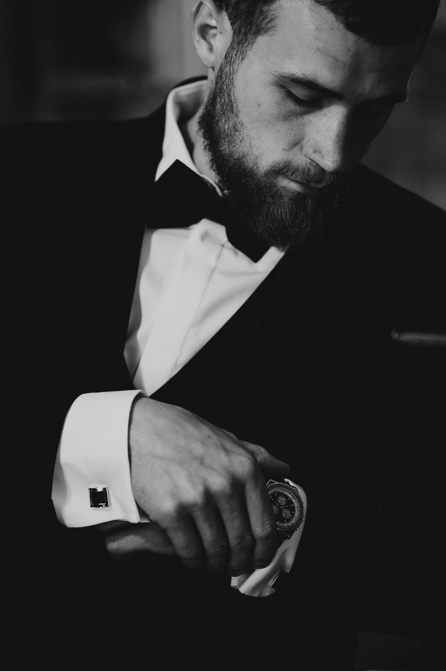 Black and white portrait of a bearded groom in a tuxedo adjusting his cufflinks and watch.