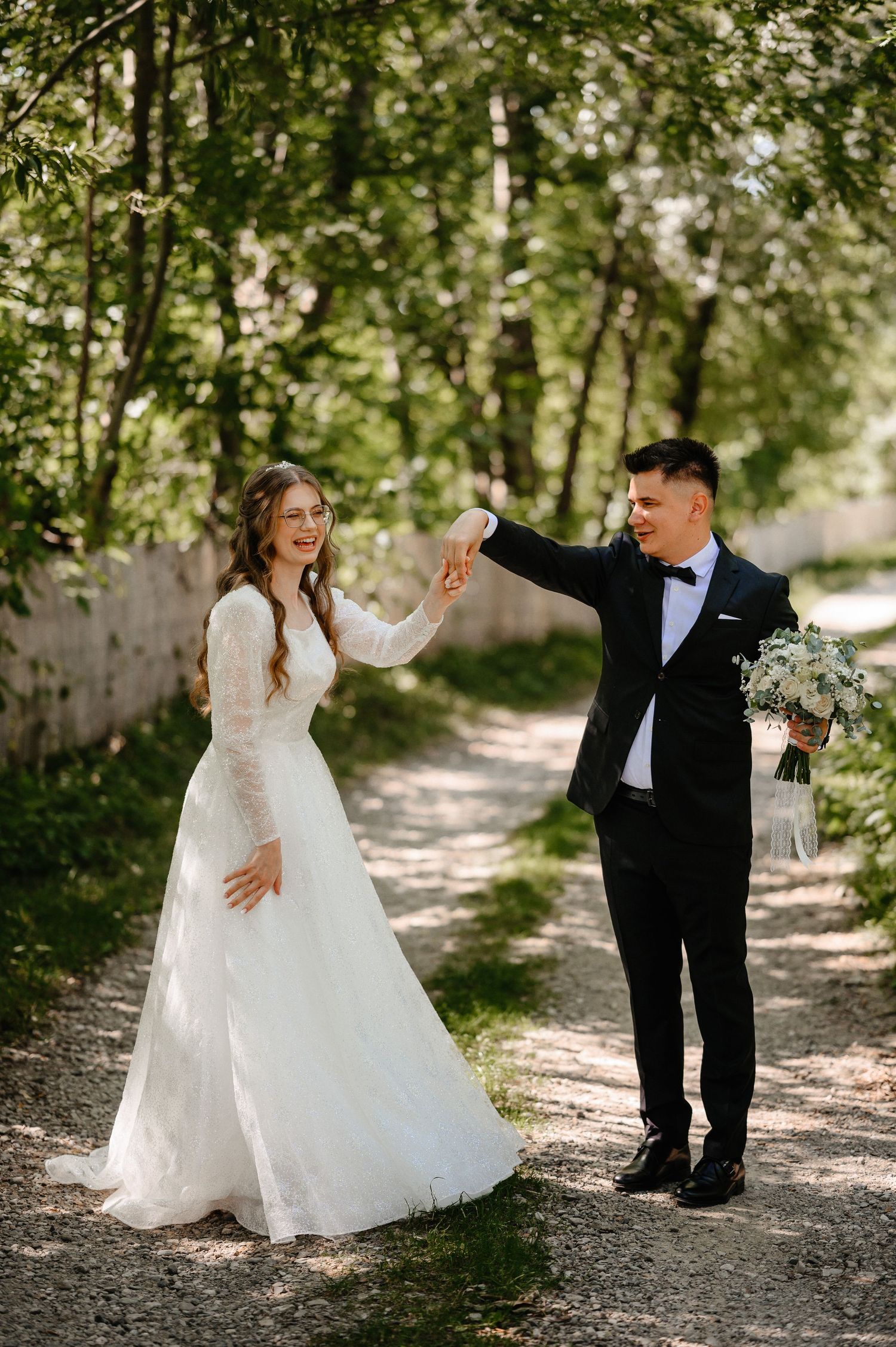 Bride in lace gown and groom in tuxedo dancing on a sunlit forest path.