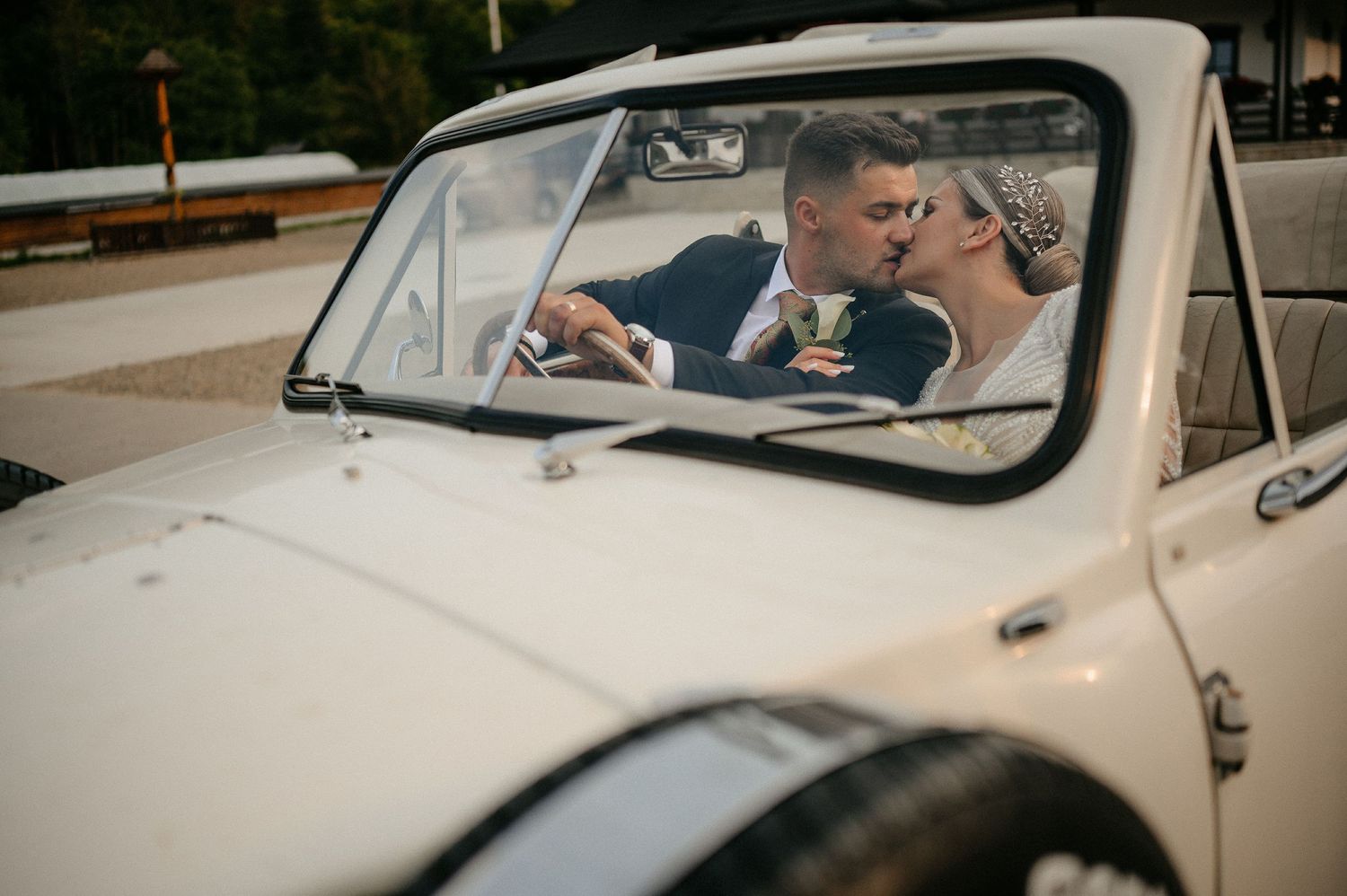 Bride and groom sharing a romantic kiss inside a classic white vintage wedding car.