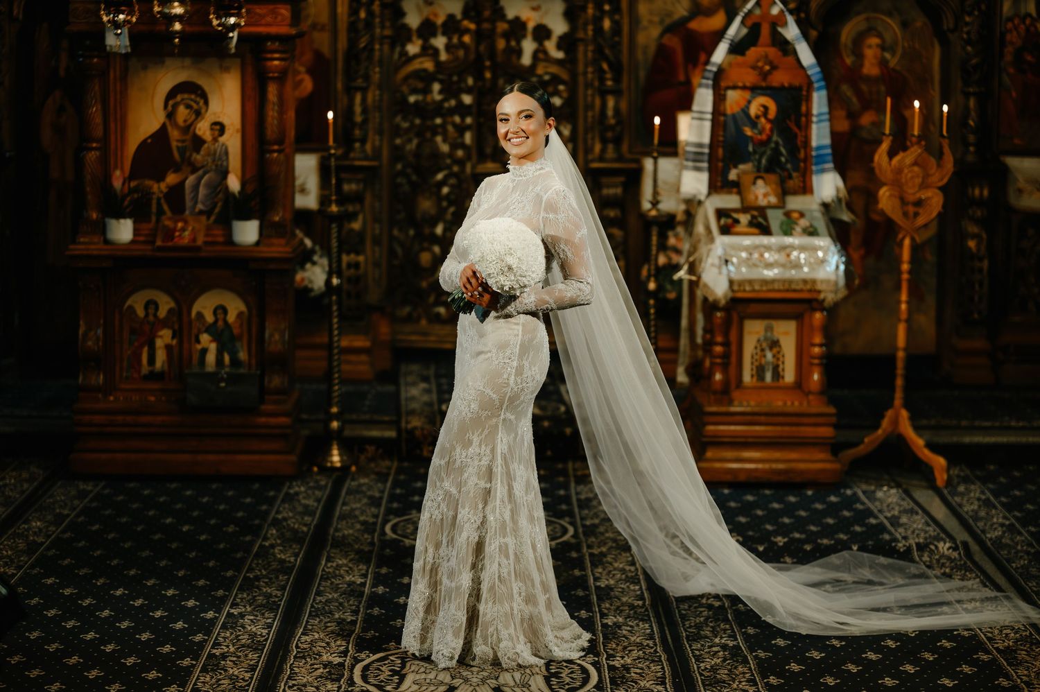 Elegant bride in a fitted lace gown holding a white bouquet standing before Orthodox church icons.