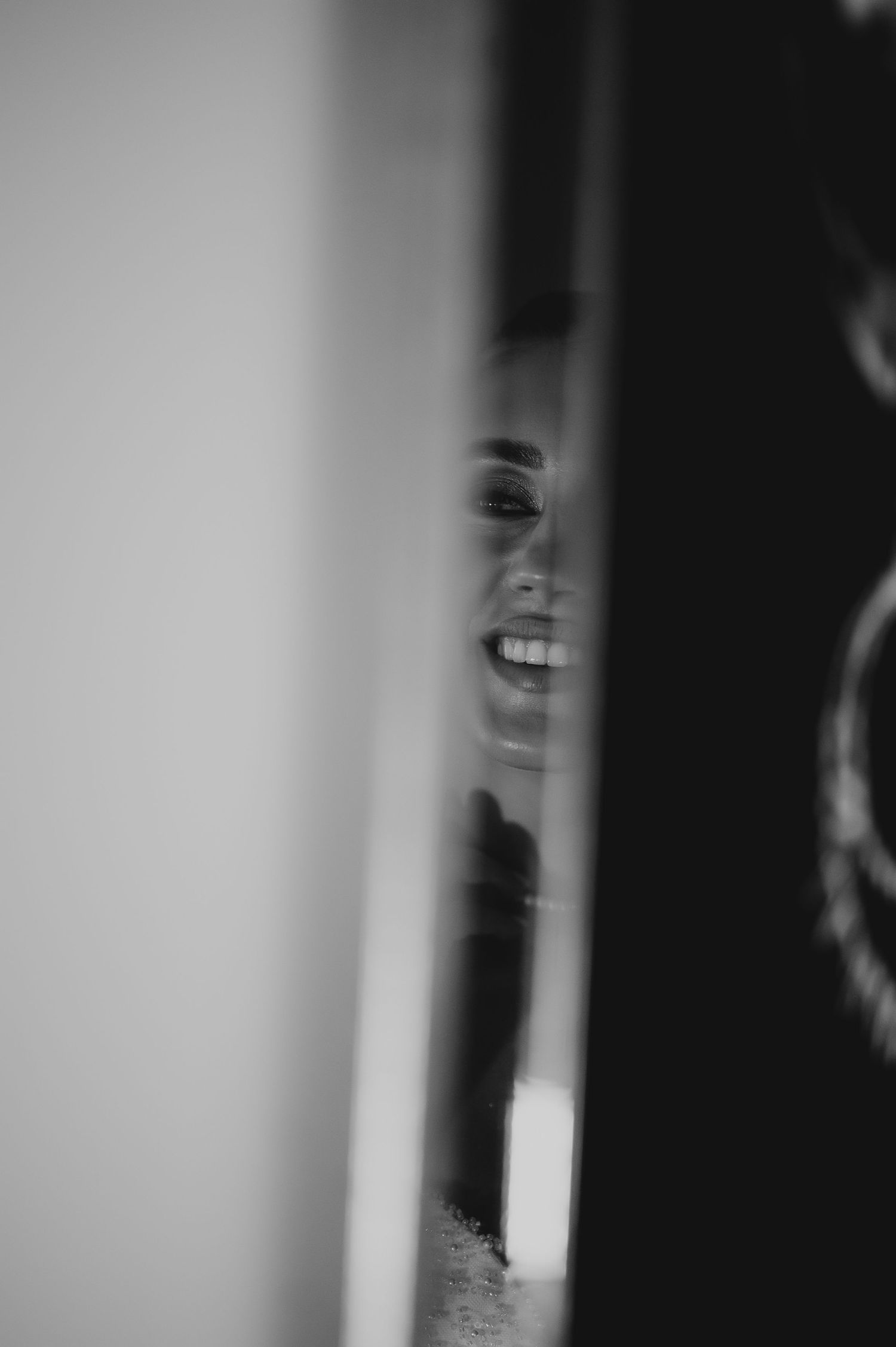 Black and white candid photo of smiling bride seen through narrow door gap.