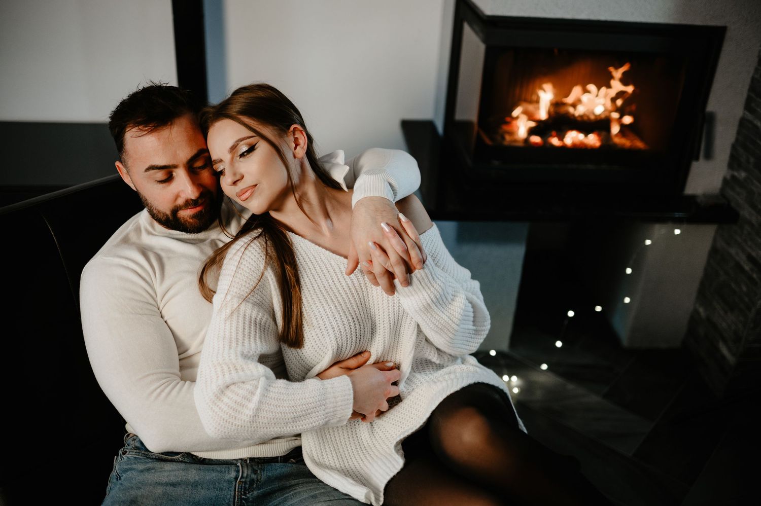 Couple in white knitwear cuddle tenderly by a warm fireplace with soft fairy lights.