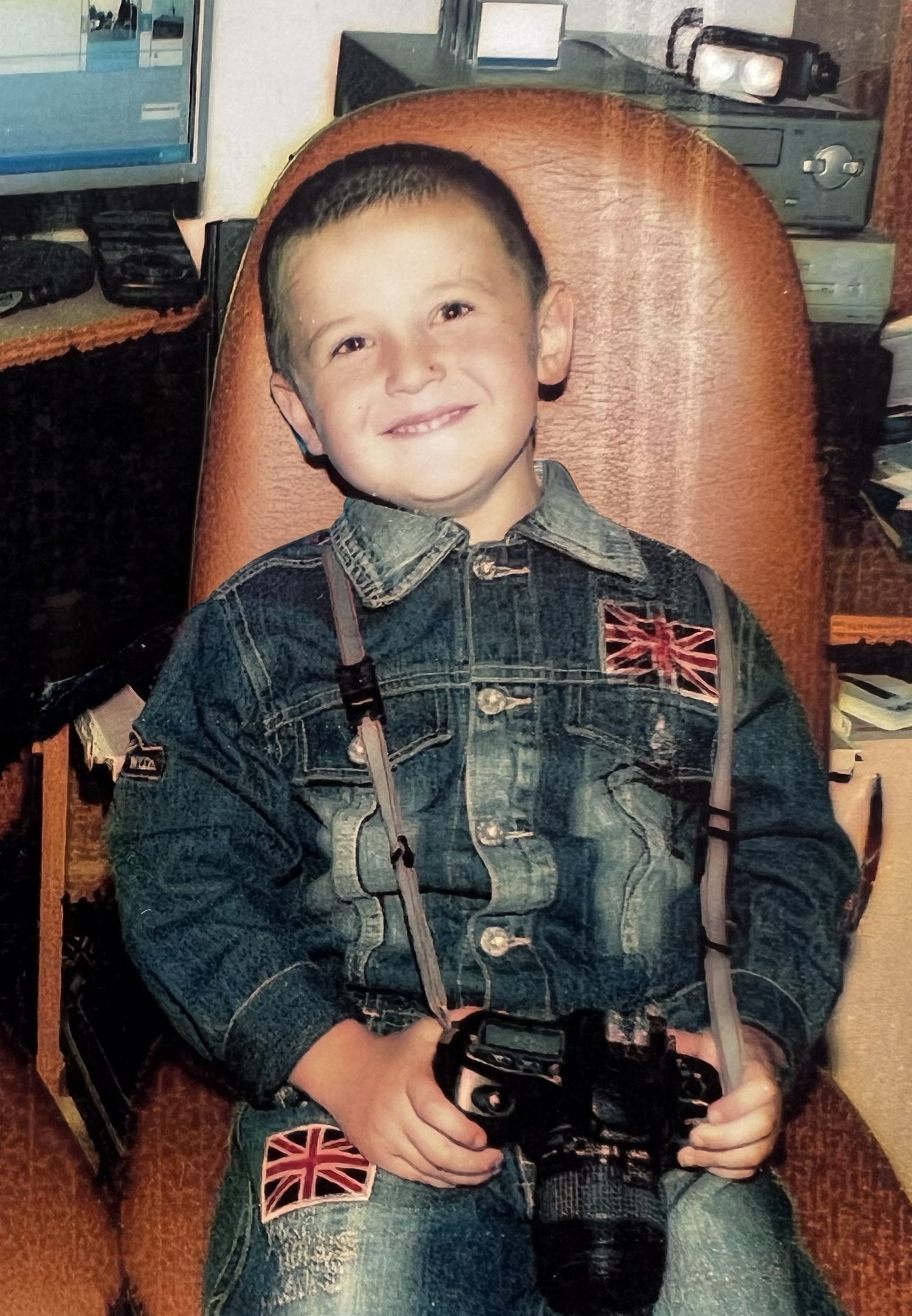 Happy young boy wearing denim Union Jack outfit holding camera while seated in brown leather chair.