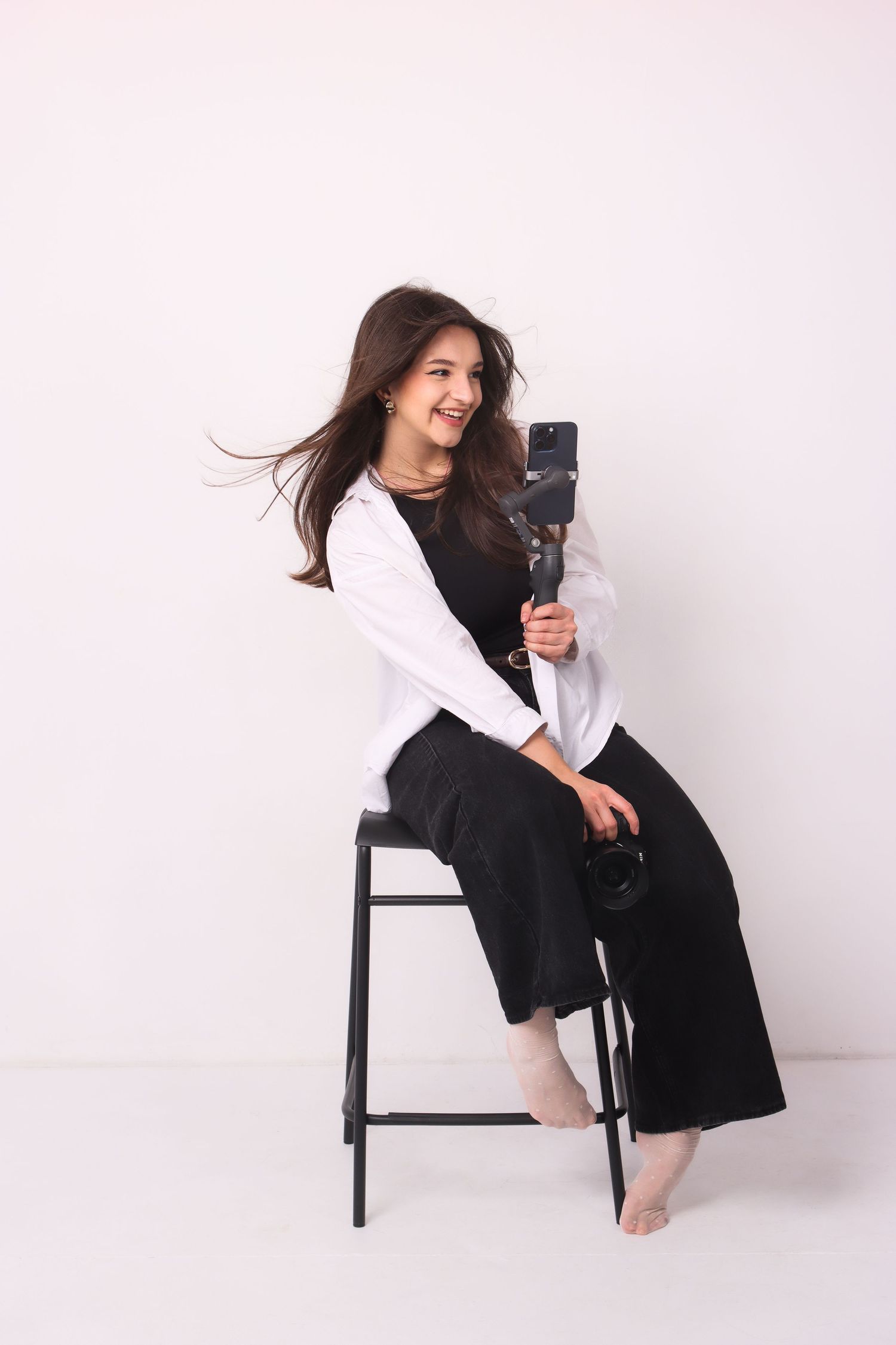 A smiling woman with flowing hair sits on stool holding smartphone gimbal on white background.