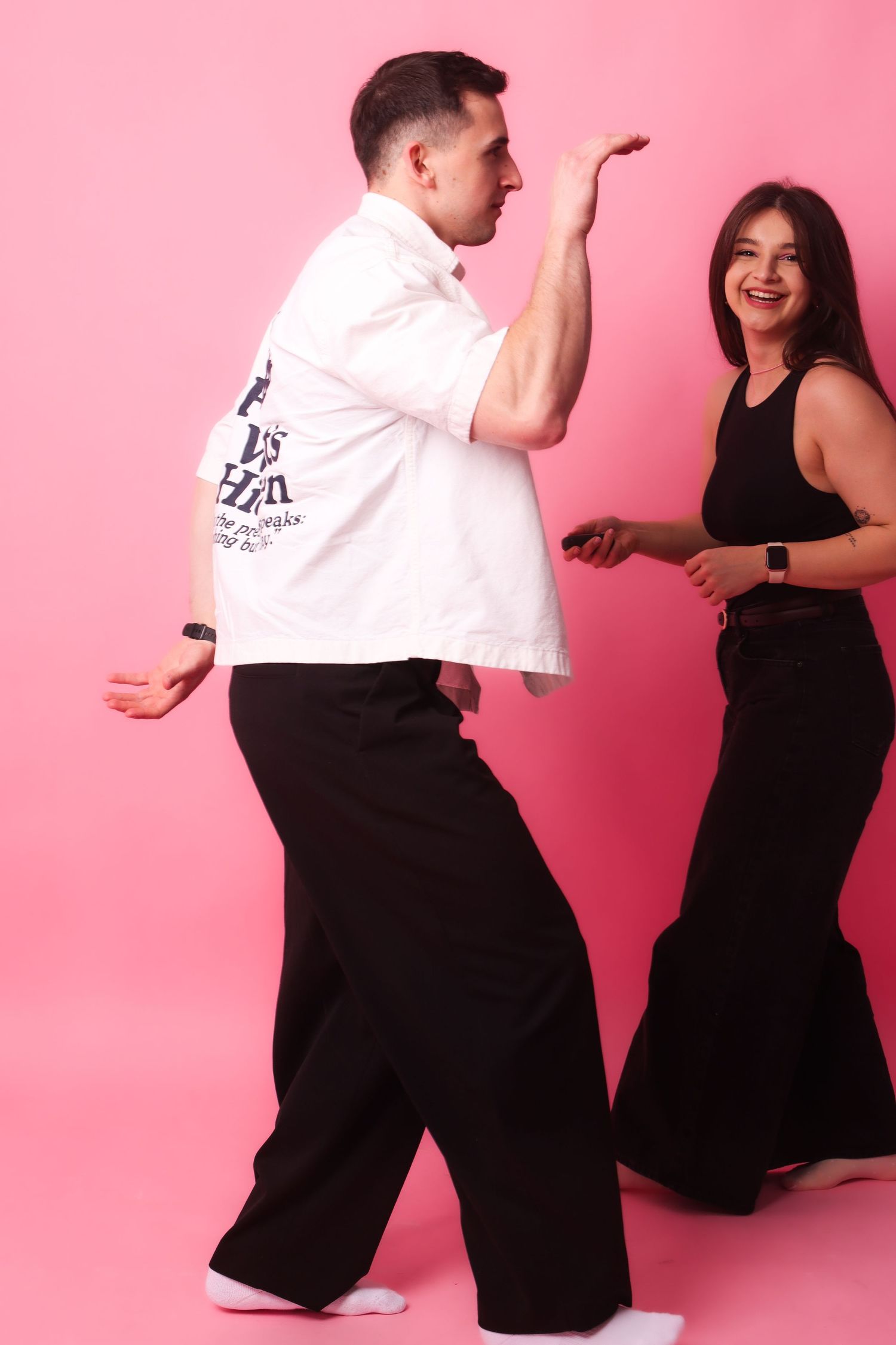 Couple dancing together in a pink studio, man in white shirt and woman in black dress smiling.
