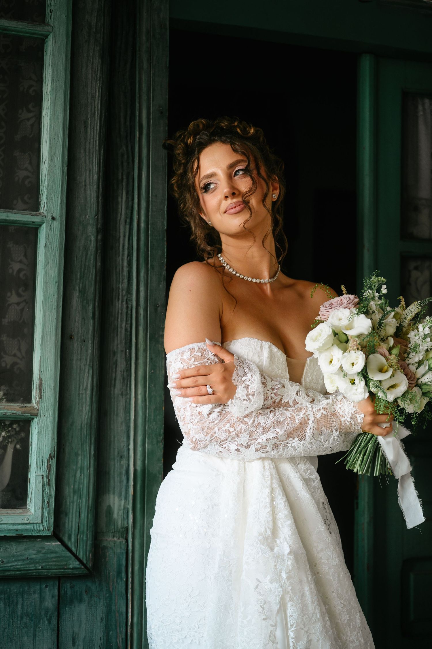 Elegant bride in lace off-shoulder gown holding white bouquet, posing by vintage green doors with pearl necklace.