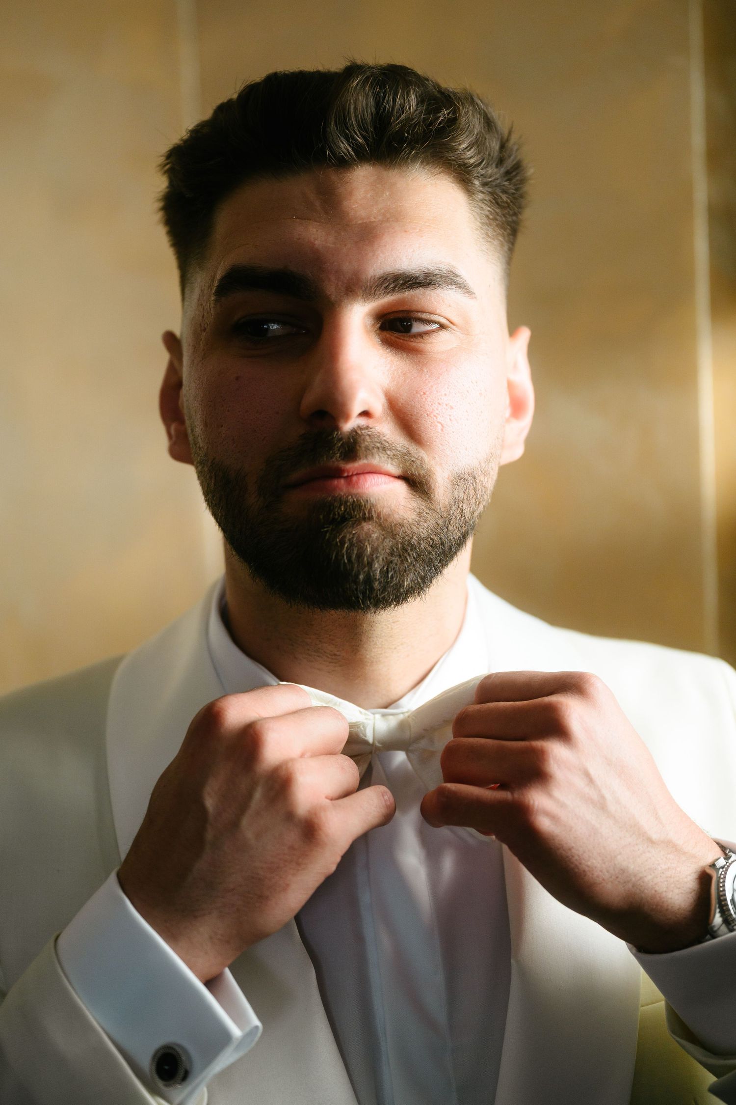 A well-dressed man in a white tuxedo adjusting his bow tie, wearing cufflinks and a watch.