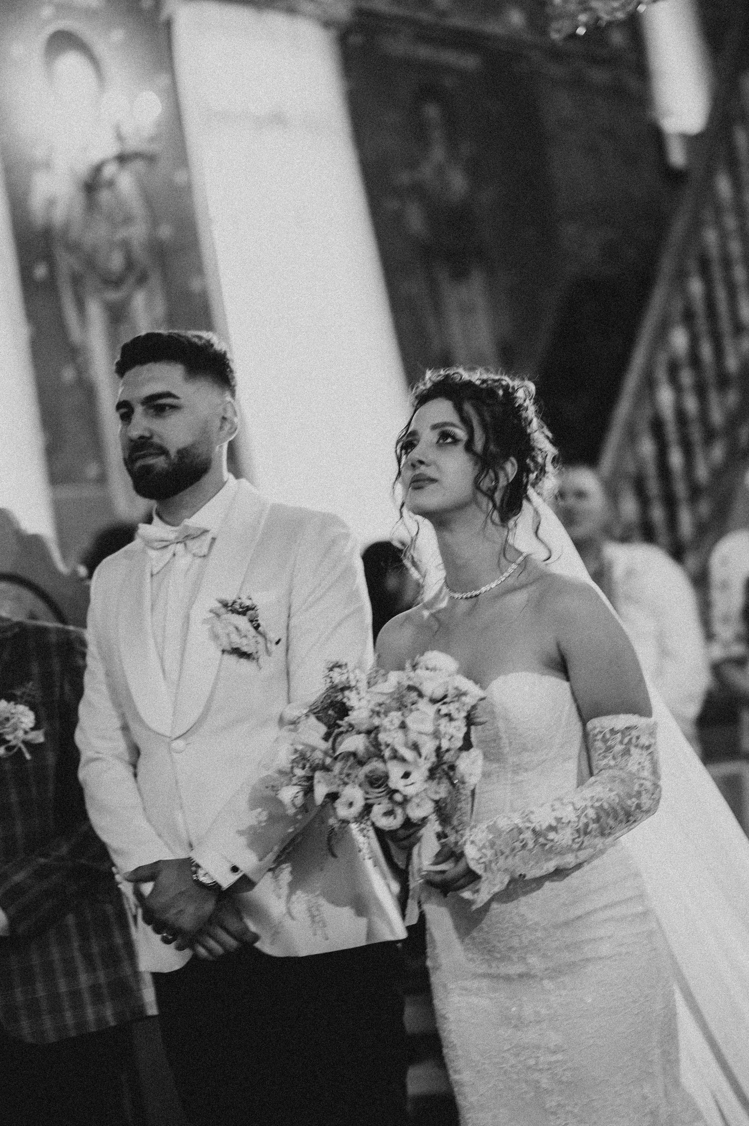 Black and white wedding photo of bride holding bouquet and groom in white tuxedo standing at church altar.