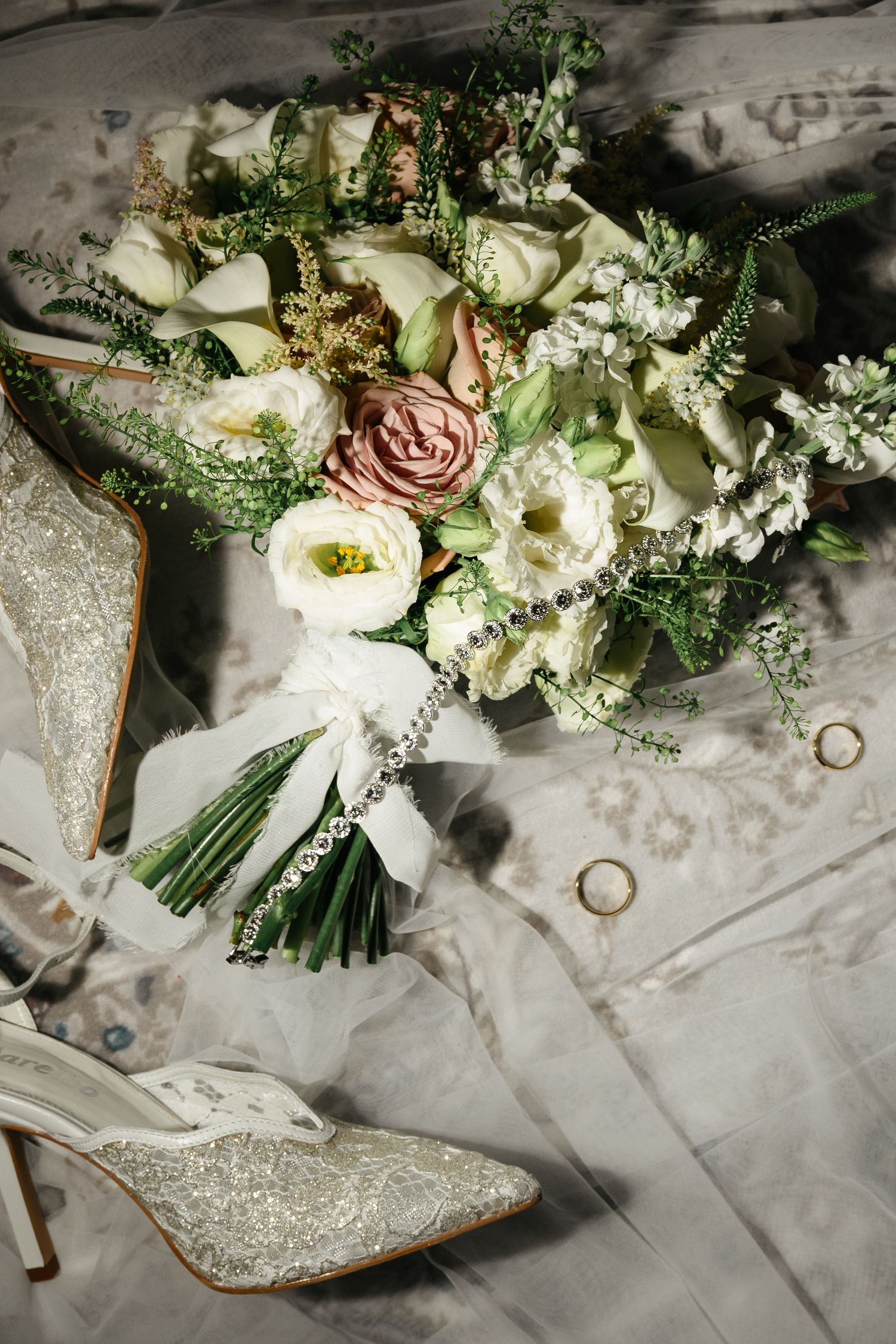 Elegant bridal flat lay featuring a lush white and pink bouquet, sparkly heels, wedding rings, and a delicate veil.