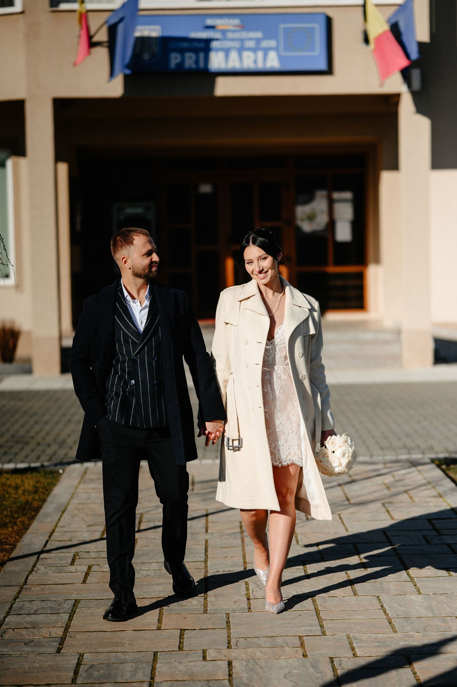 Newlywed couple holding hands and smiling outside a Romanian Primaria building, bride in white coat holding bouquet.