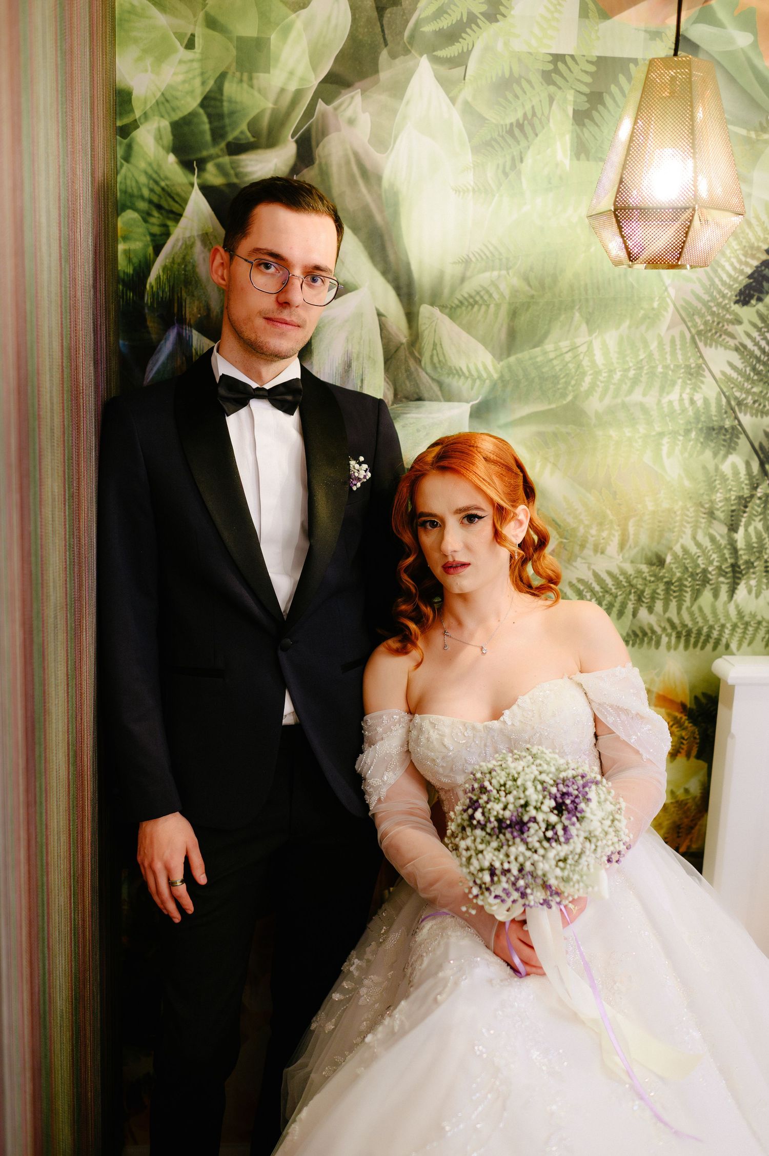 Bride in white off-shoulder gown holding bouquet stands with groom in black tuxedo before green botanical wallpaper.