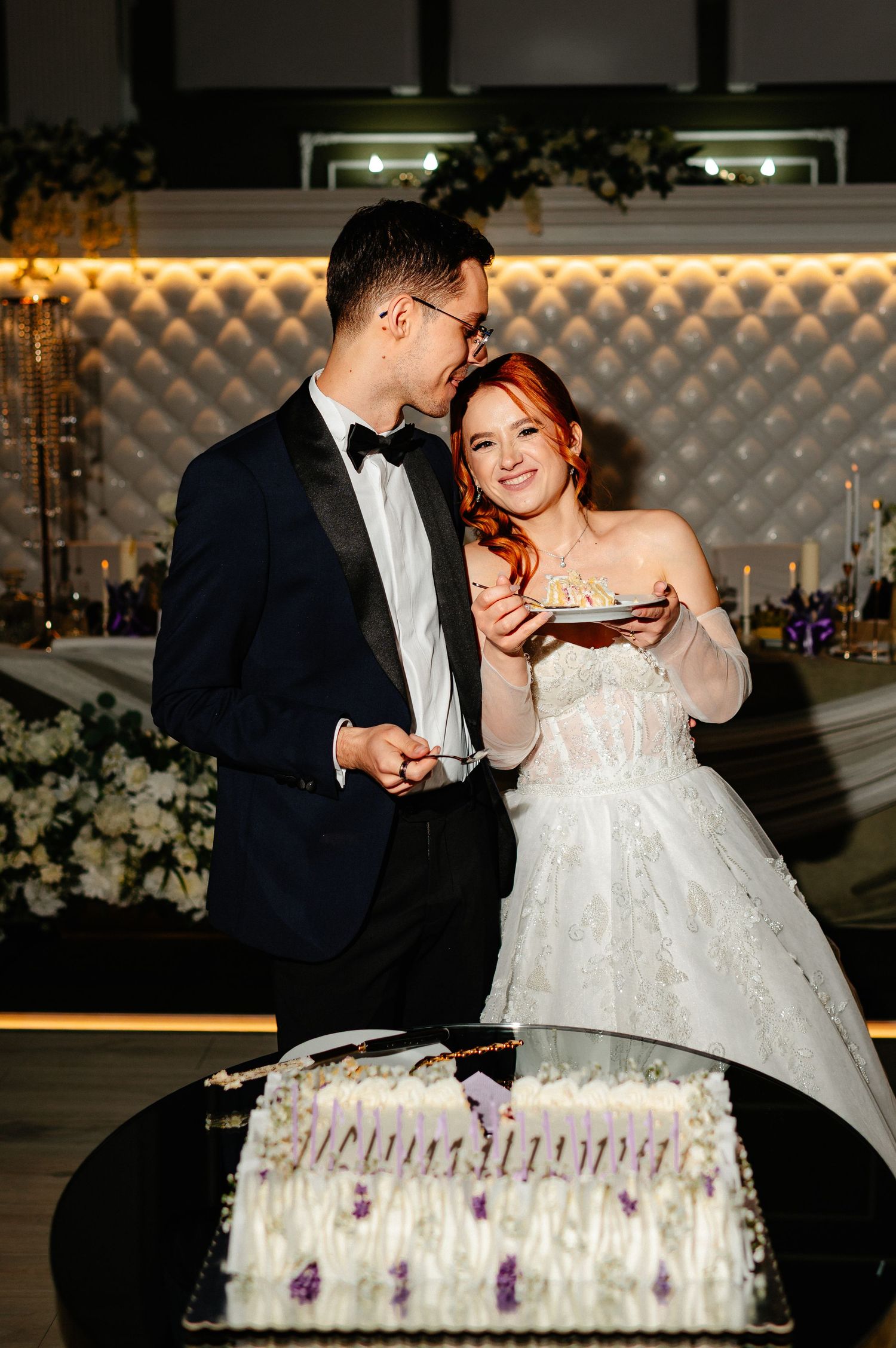 Bride and groom cutting white wedding cake at elegant reception, bride in strapless gown smiling, groom in black tuxedo.