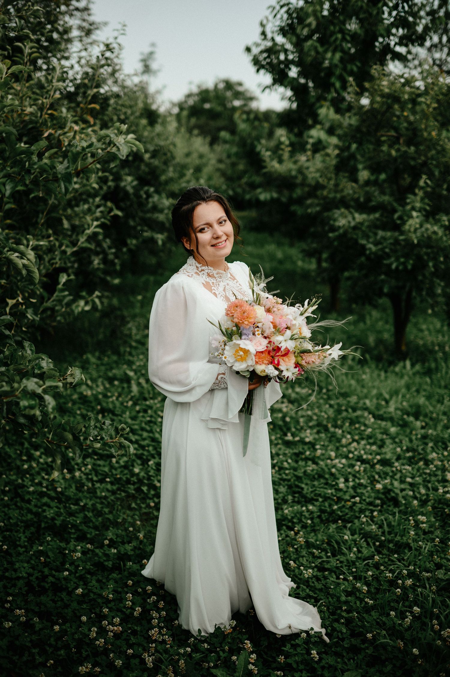 Bride in white gown holding colorful bouquet standing in lush green orchard smiling on her wedding day.