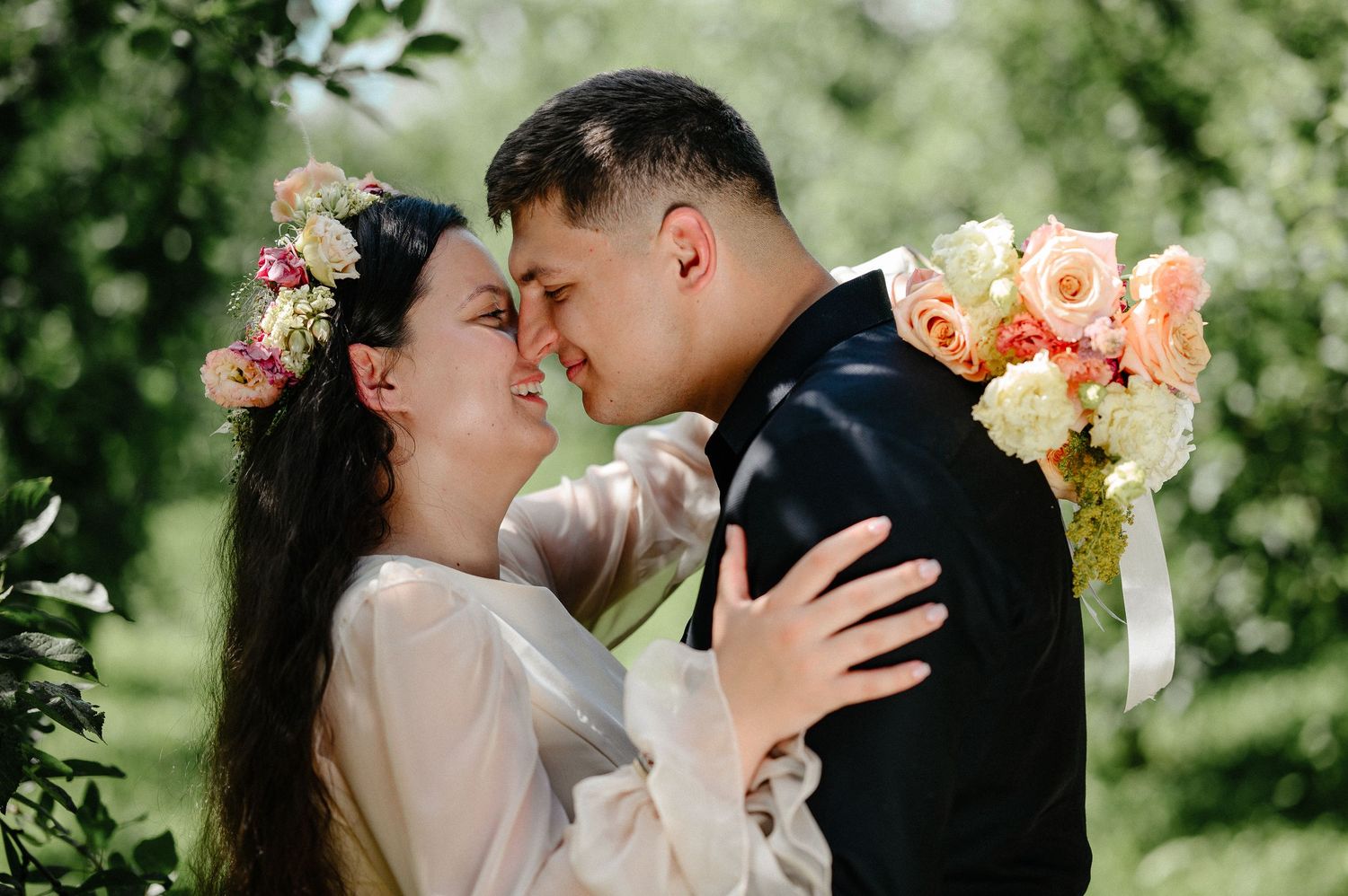 A bride and groom share a romantic moment outdoors, with floral crown and bouquet, surrounded by lush green trees.