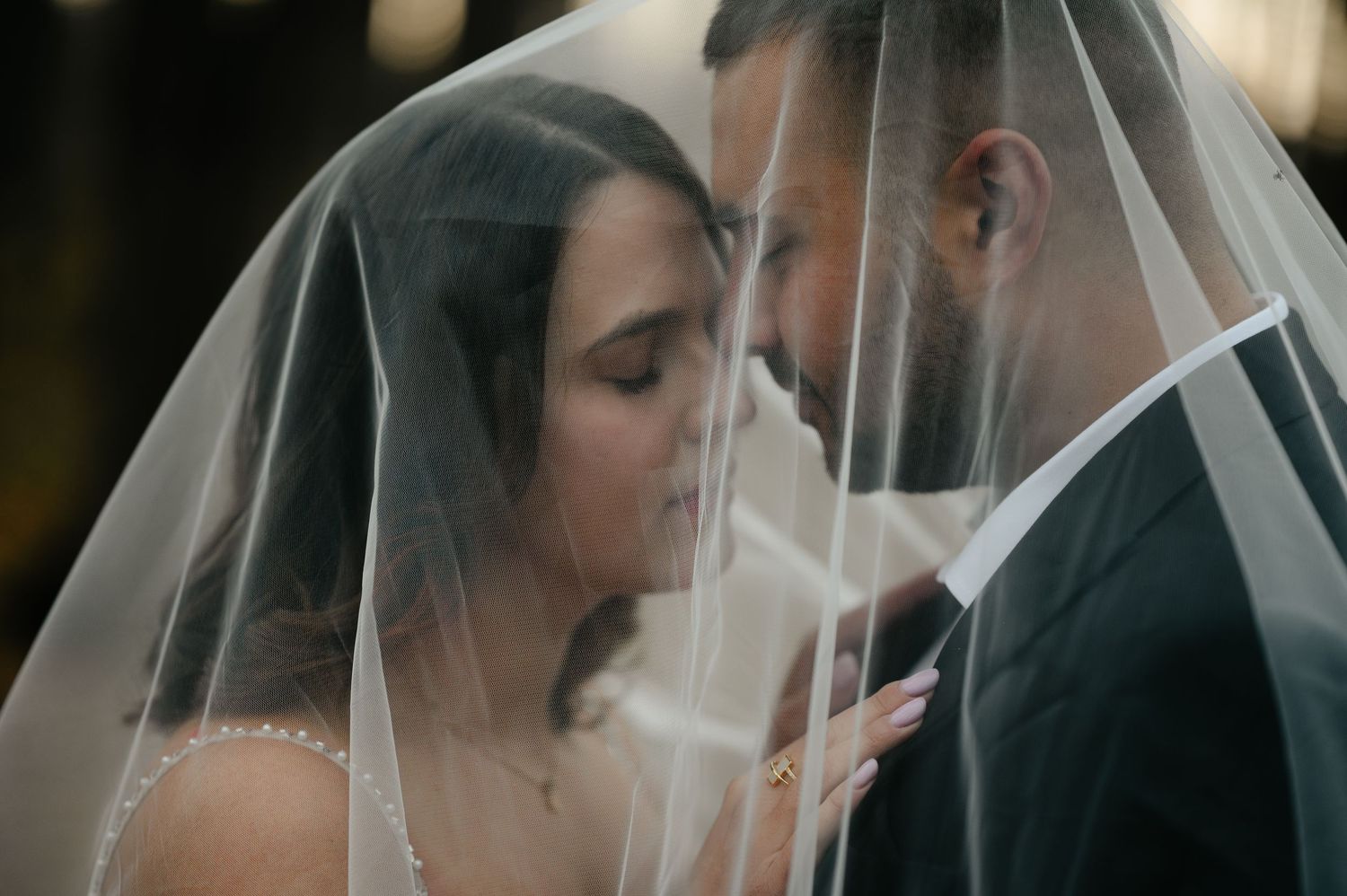 Bride and groom share an intimate moment under a sheer wedding veil, foreheads nearly touching in a romantic pose.