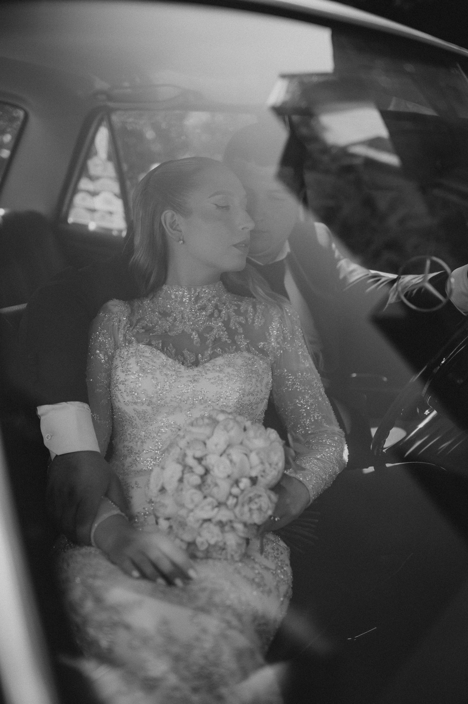Black and white photo of a bride in lace gown holding bouquet, sharing intimate moment with groom in car.