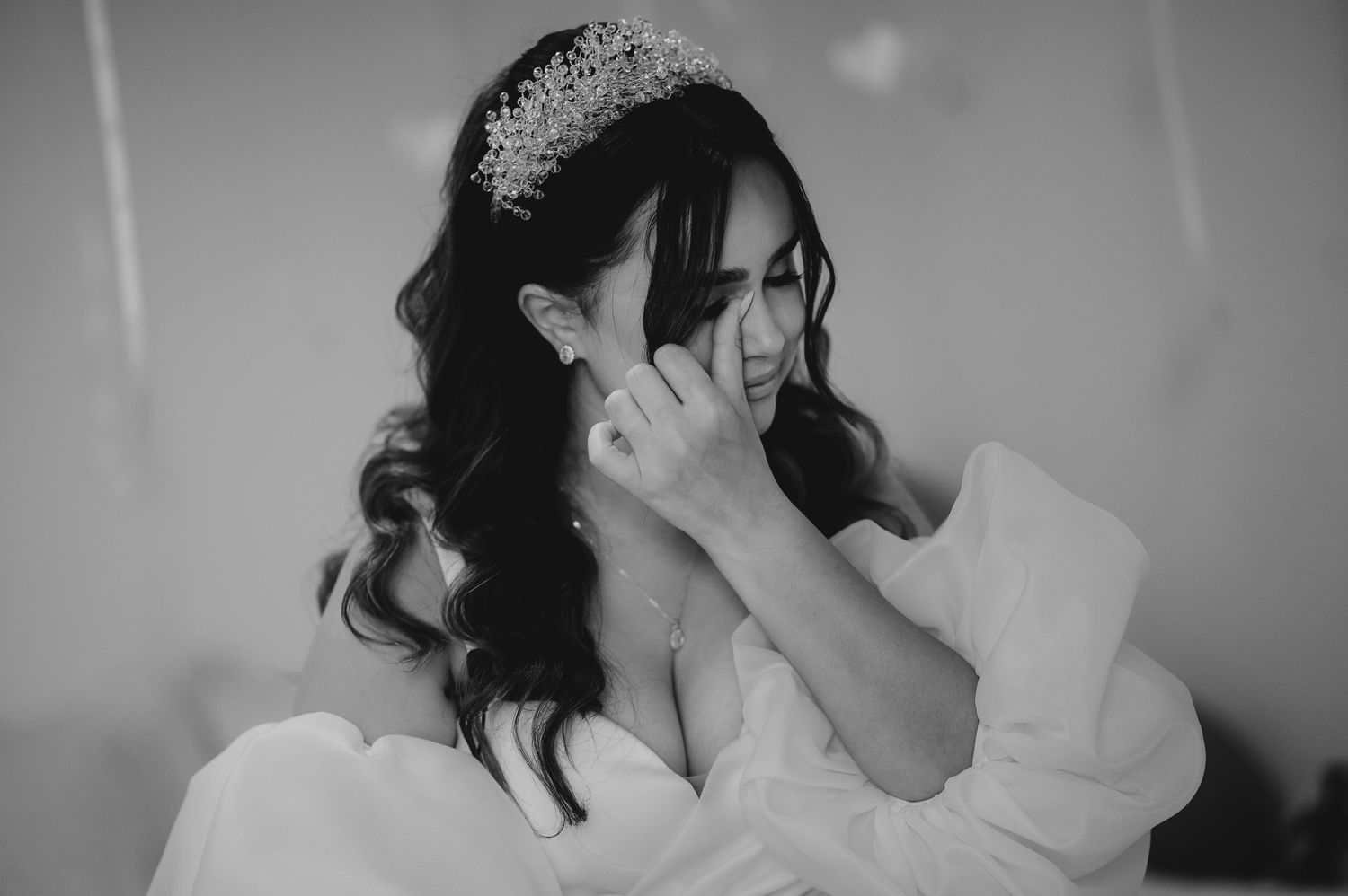 Emotional bride in white robe wearing tiara wipes tears in black and white wedding preparation photo.