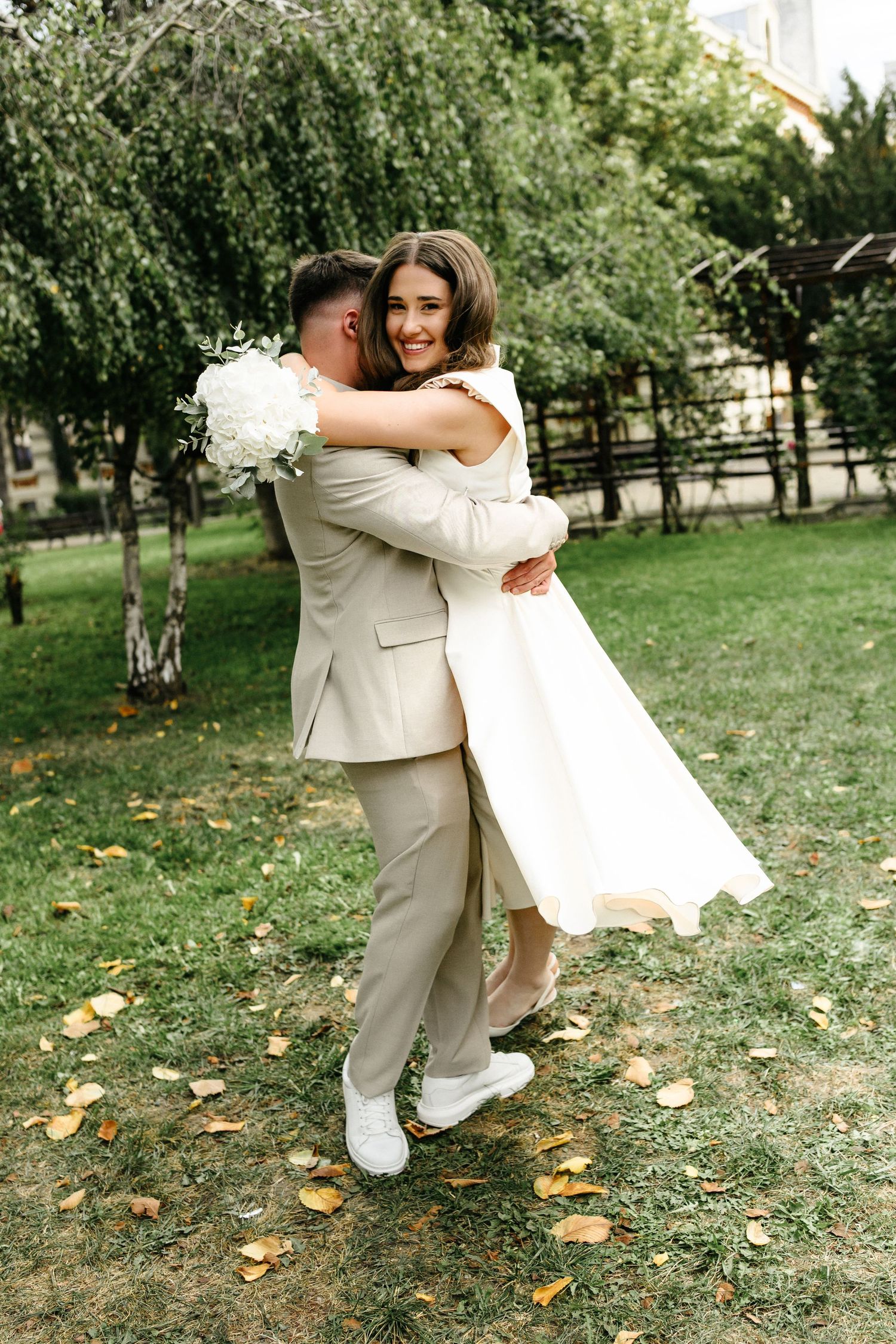 Joyful bride in white dress lifted by groom in beige suit outdoors in a lush green park setting.