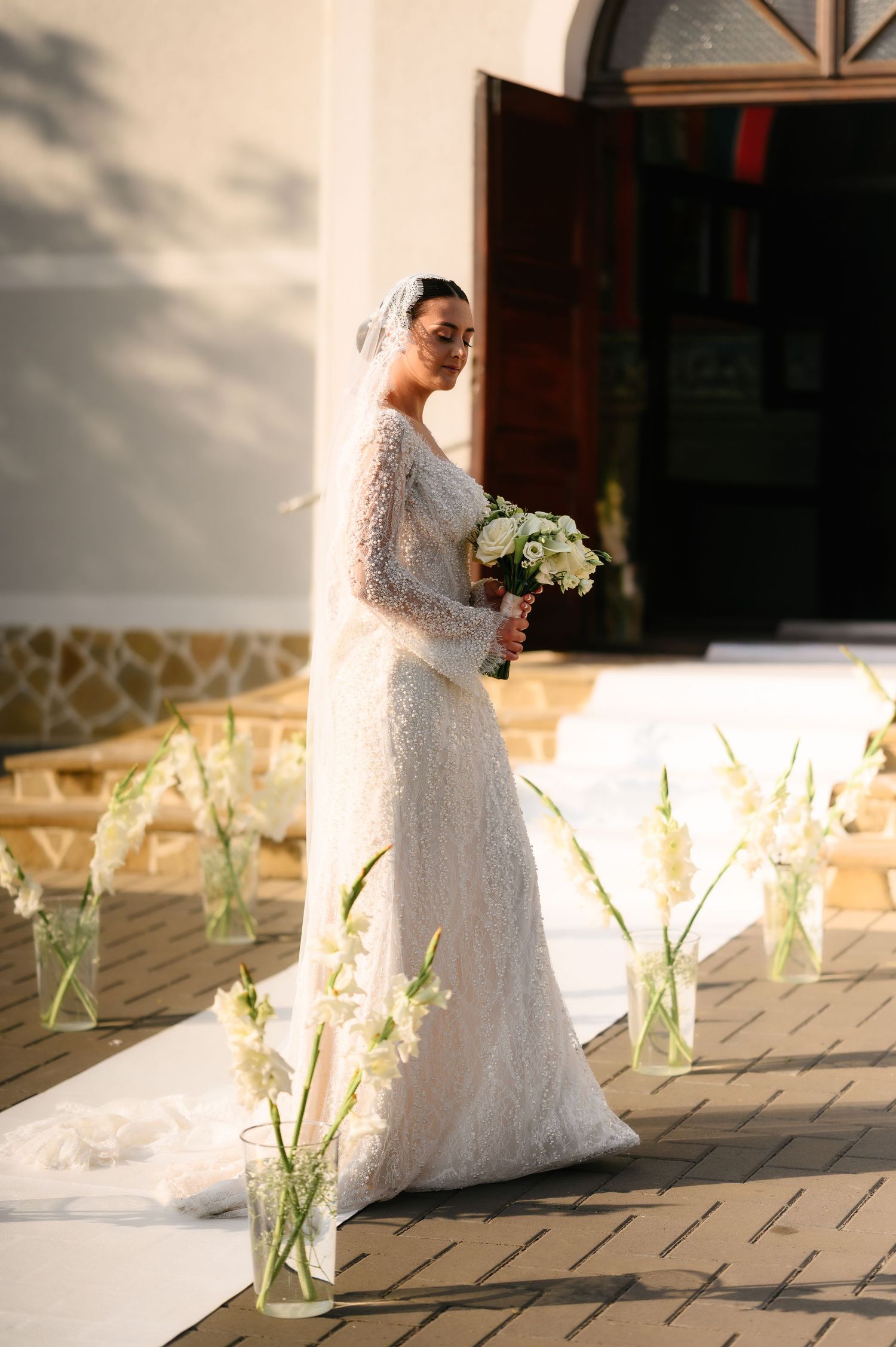 Elegant bride in long-sleeve lace wedding gown holding white bouquet, standing beside floral aisle arrangements outside a church.