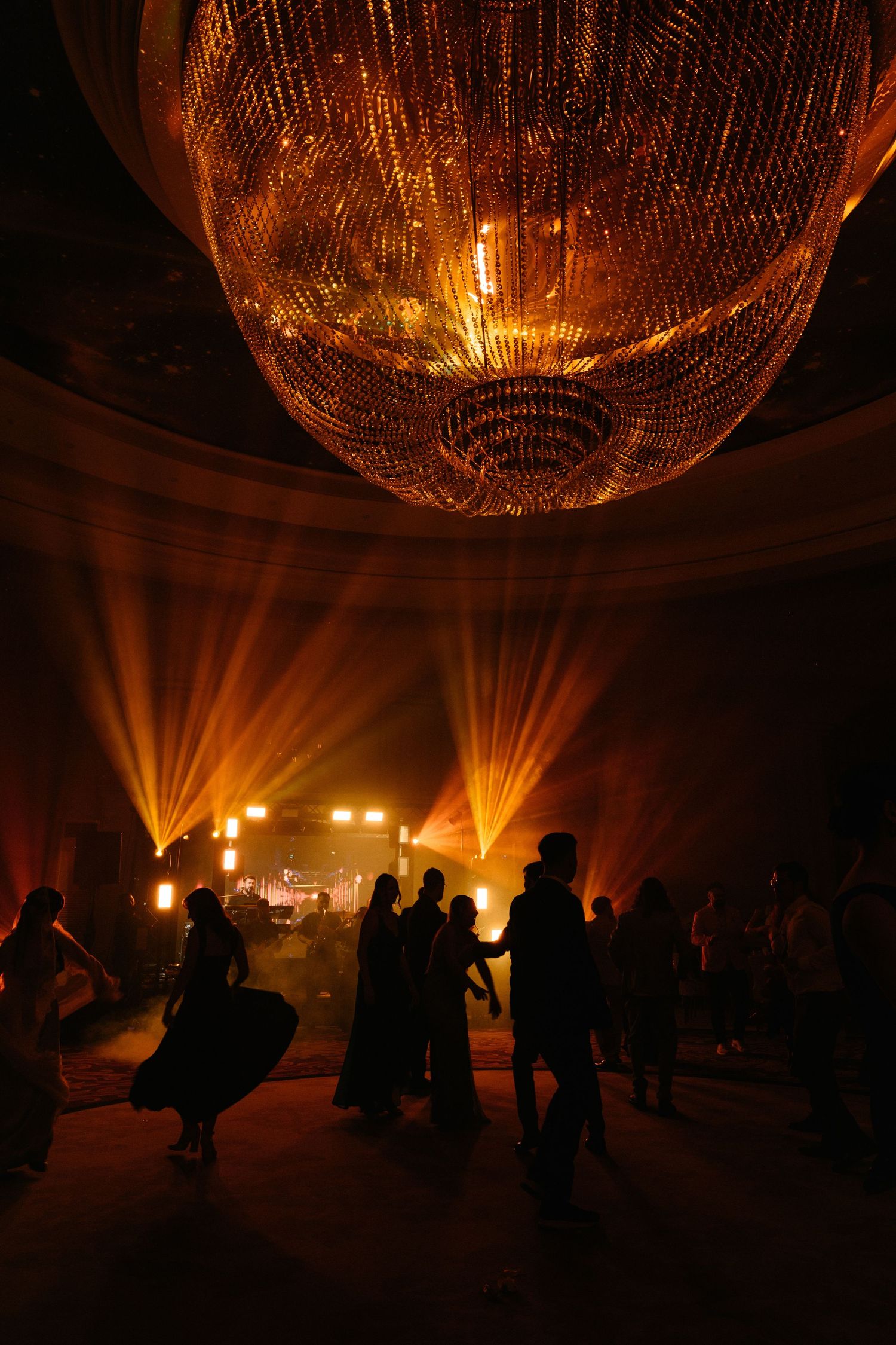 Silhouettes of dancers under a grand crystal chandelier with warm golden stage lights at an elegant event.
