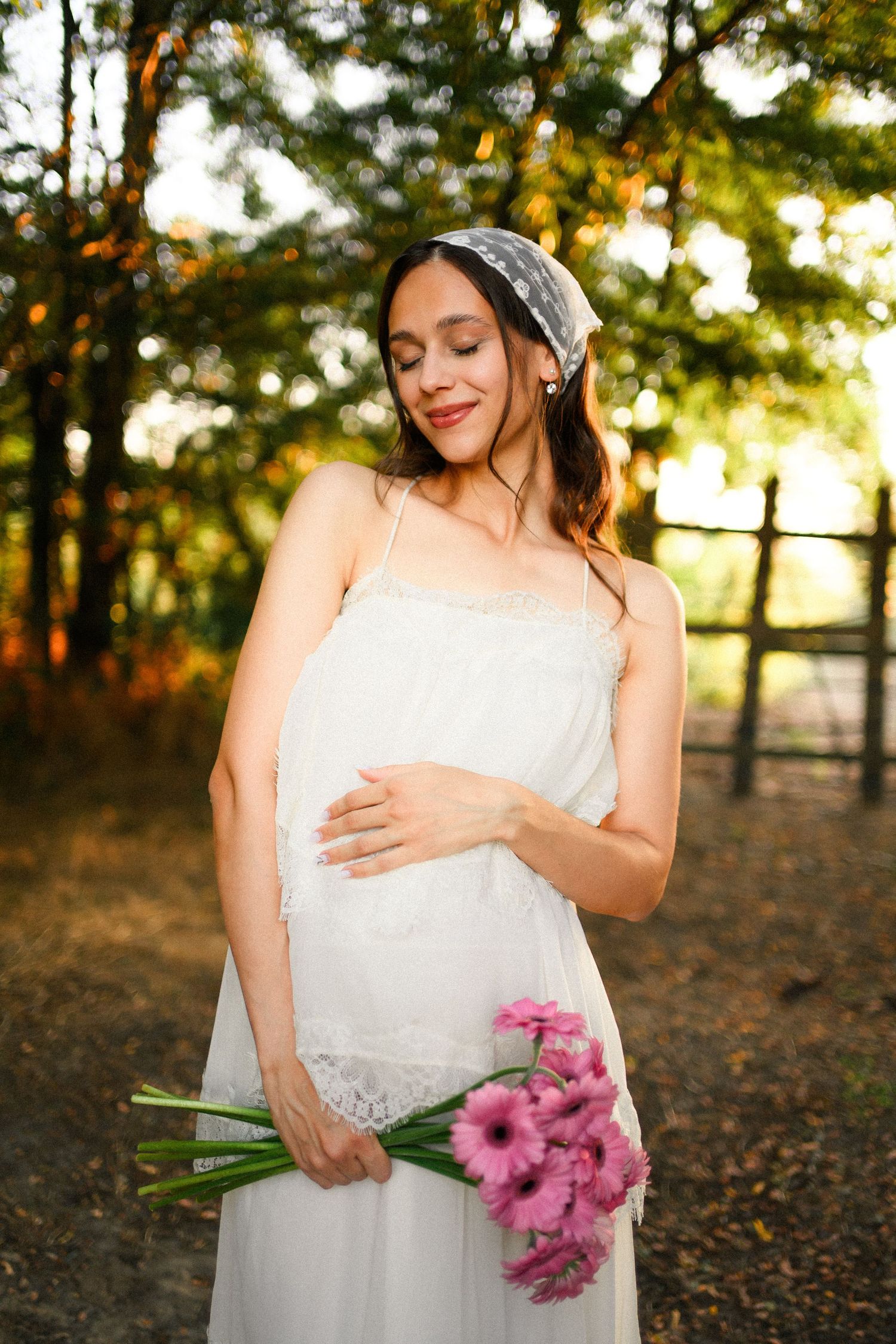 Pregnant woman in white lace dress holding pink flowers outdoors in autumn forest setting.