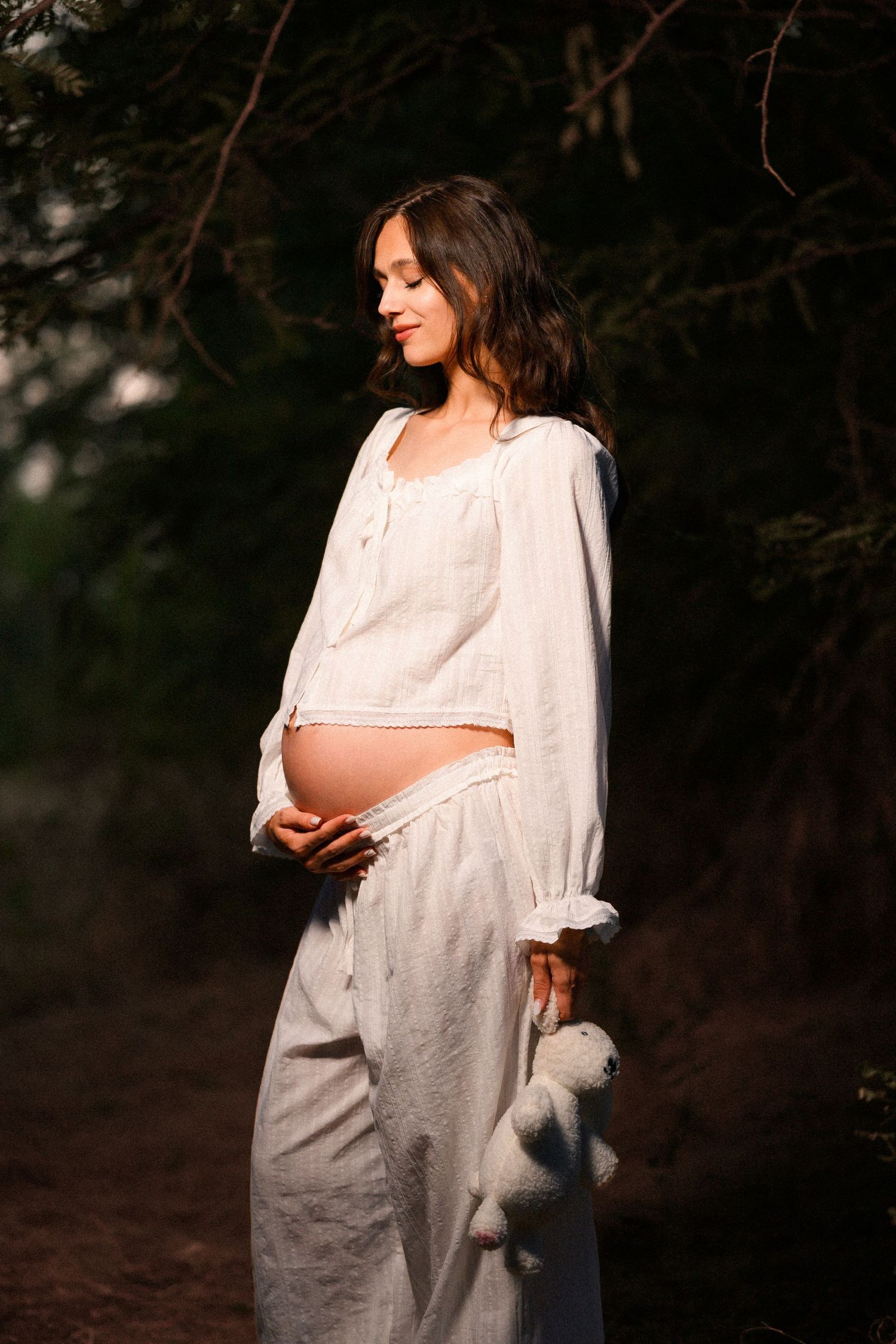 Pregnant woman in white linen outfit holding a teddy bear outdoors in soft golden light.