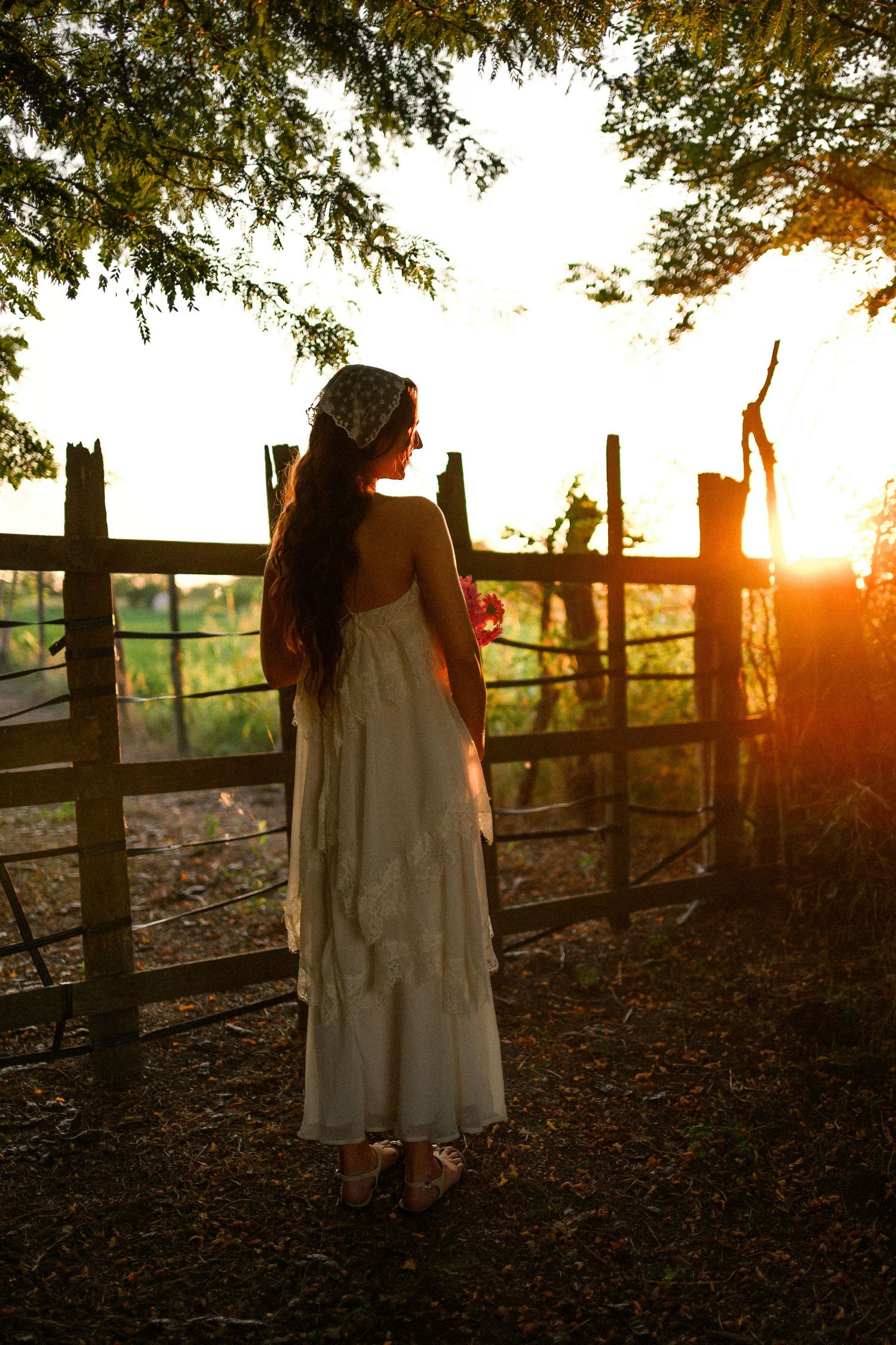 Woman in white dress and headscarf standing by rustic fence at golden sunset.