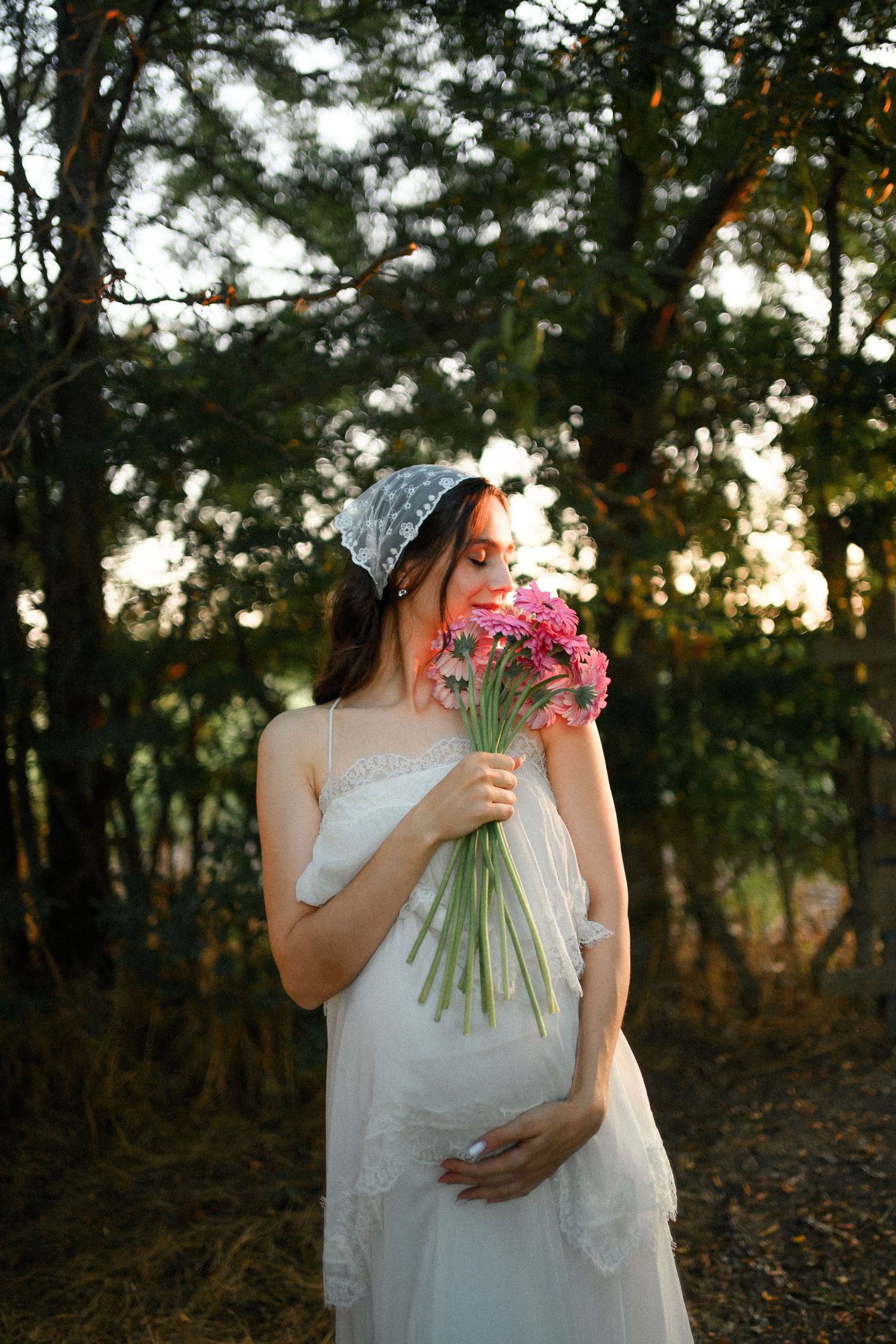 Pregnant woman in white dress smelling pink flowers in a sunlit forest, wearing a lace headscarf.