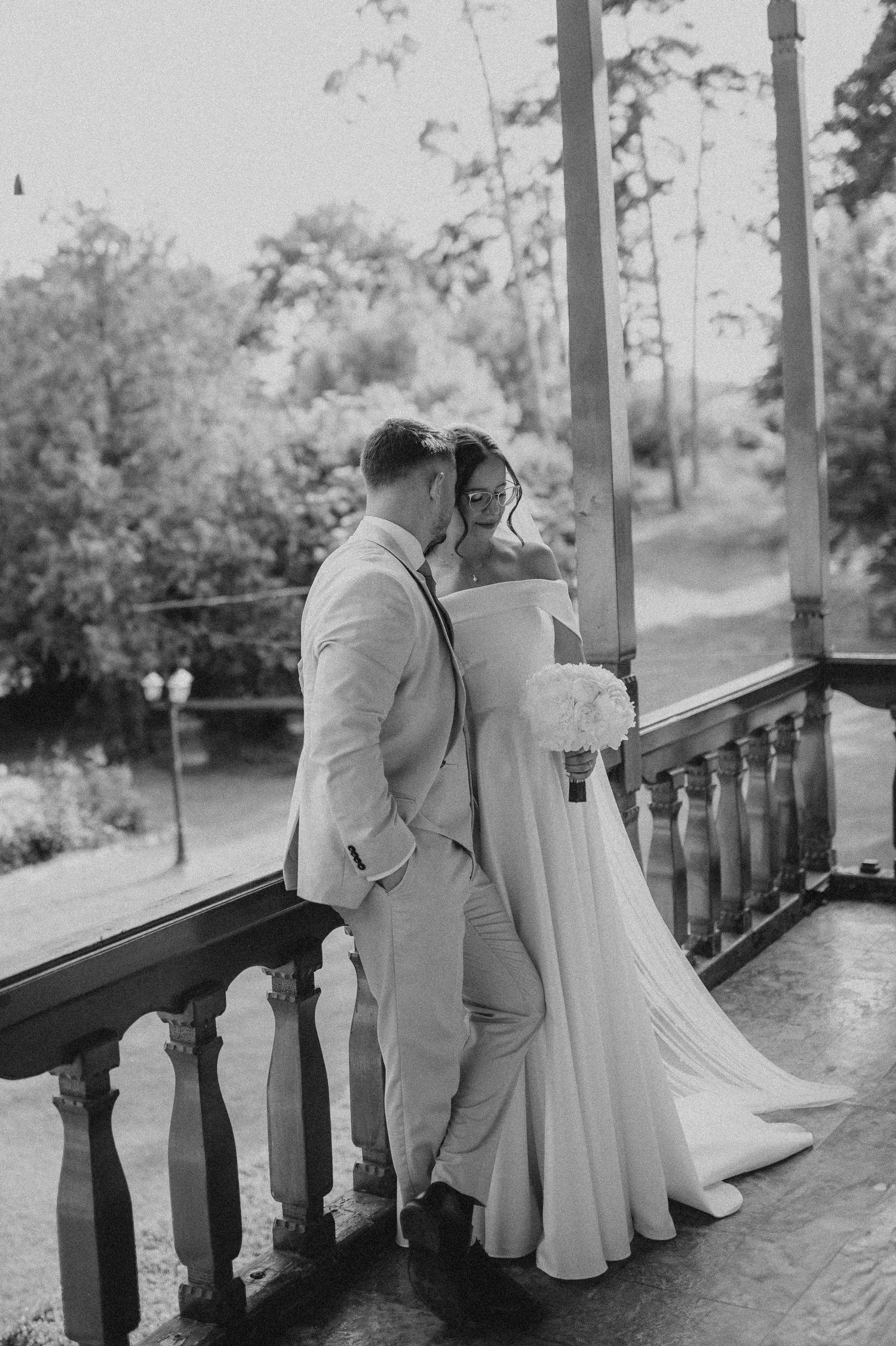 Black and white romantic photo of bride and groom embracing on outdoor balcony surrounded by trees.