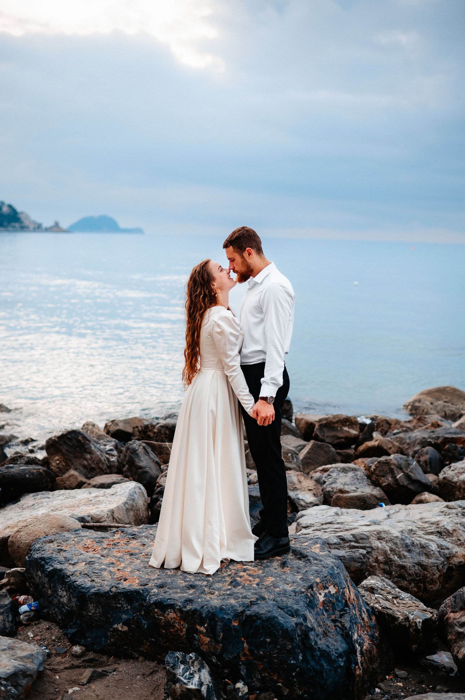 Romantic couple about to kiss while standing on rocky coastal shore at sunset.